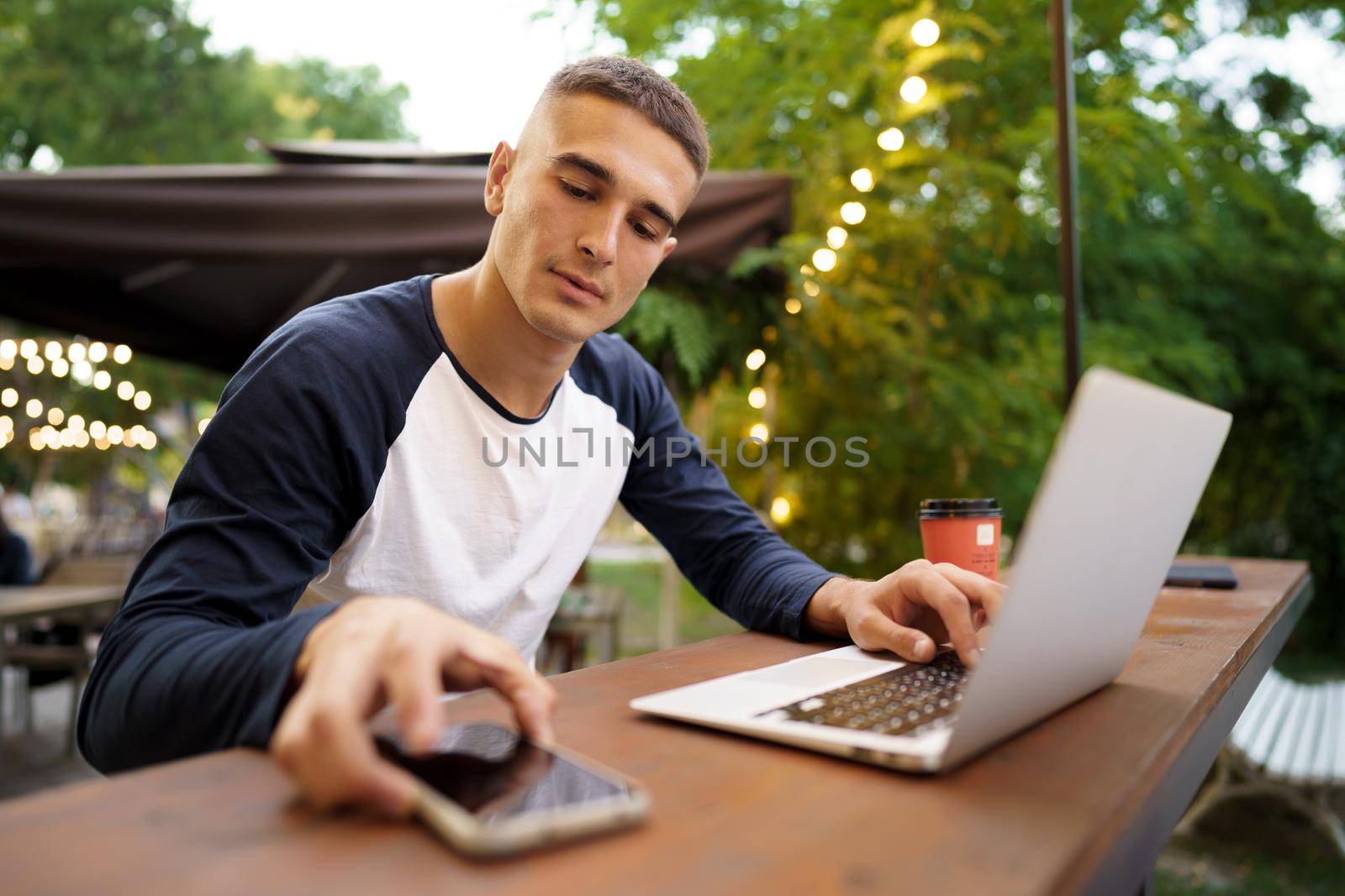 Young man sitting at table and typing on laptop keyboard while working in outdoor cafe by Fabrikasimf