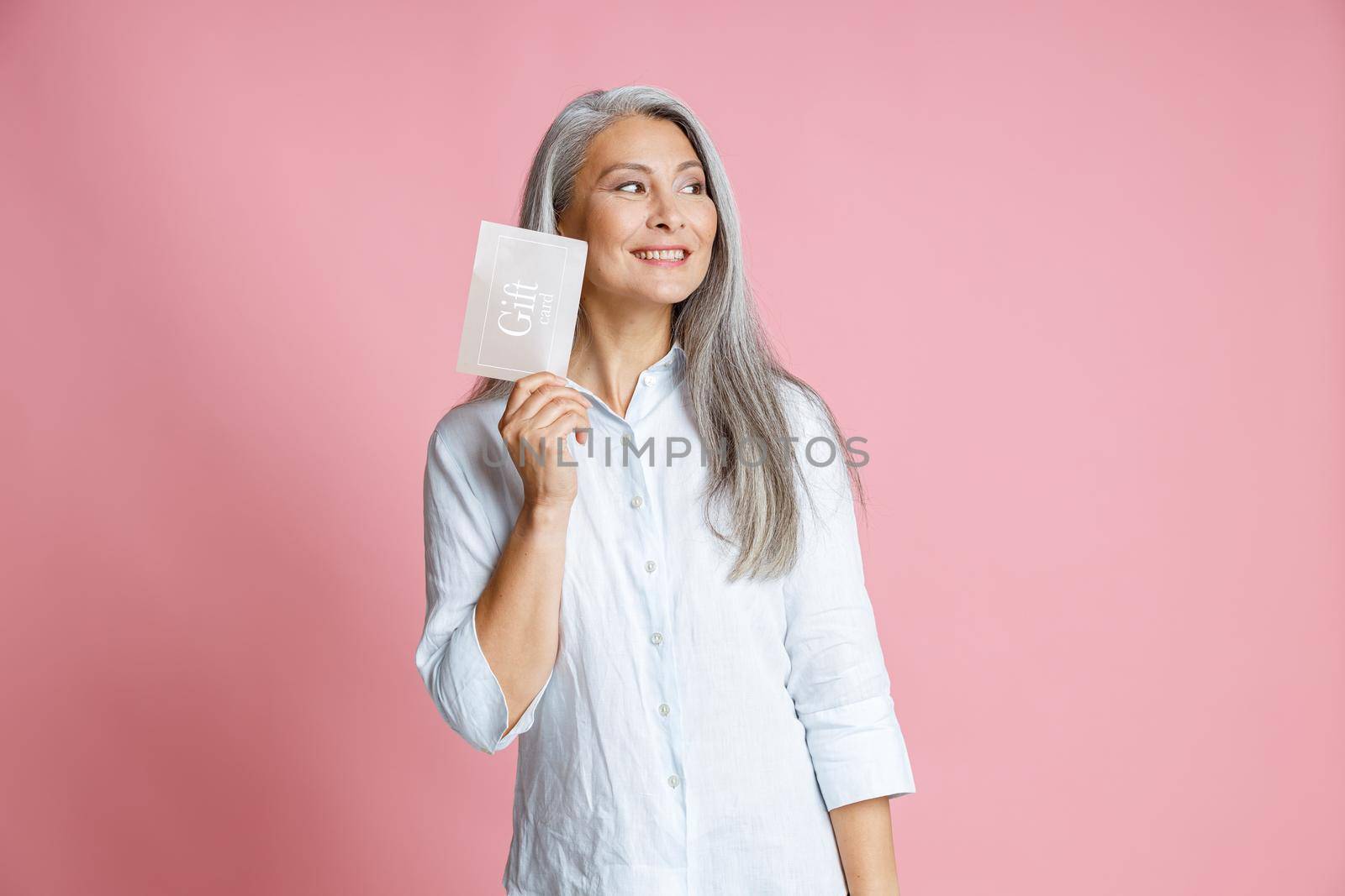 Pretty cheerful silver haired Asian woman holds gift card having ideas on pink background in studio. Shopping certificate