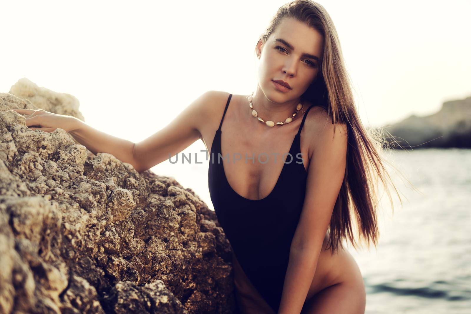 Young beautiful sexy female model in black bikini standing in sea water near huge rock