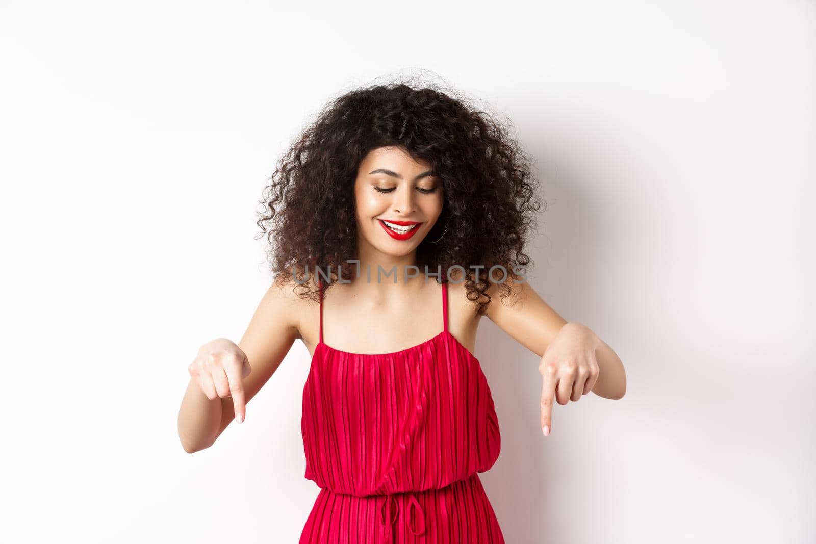 Cheerful elegant woman in red dress and makeup, looking and pointing down with pleased smile, showing advertisement, standing over white background.