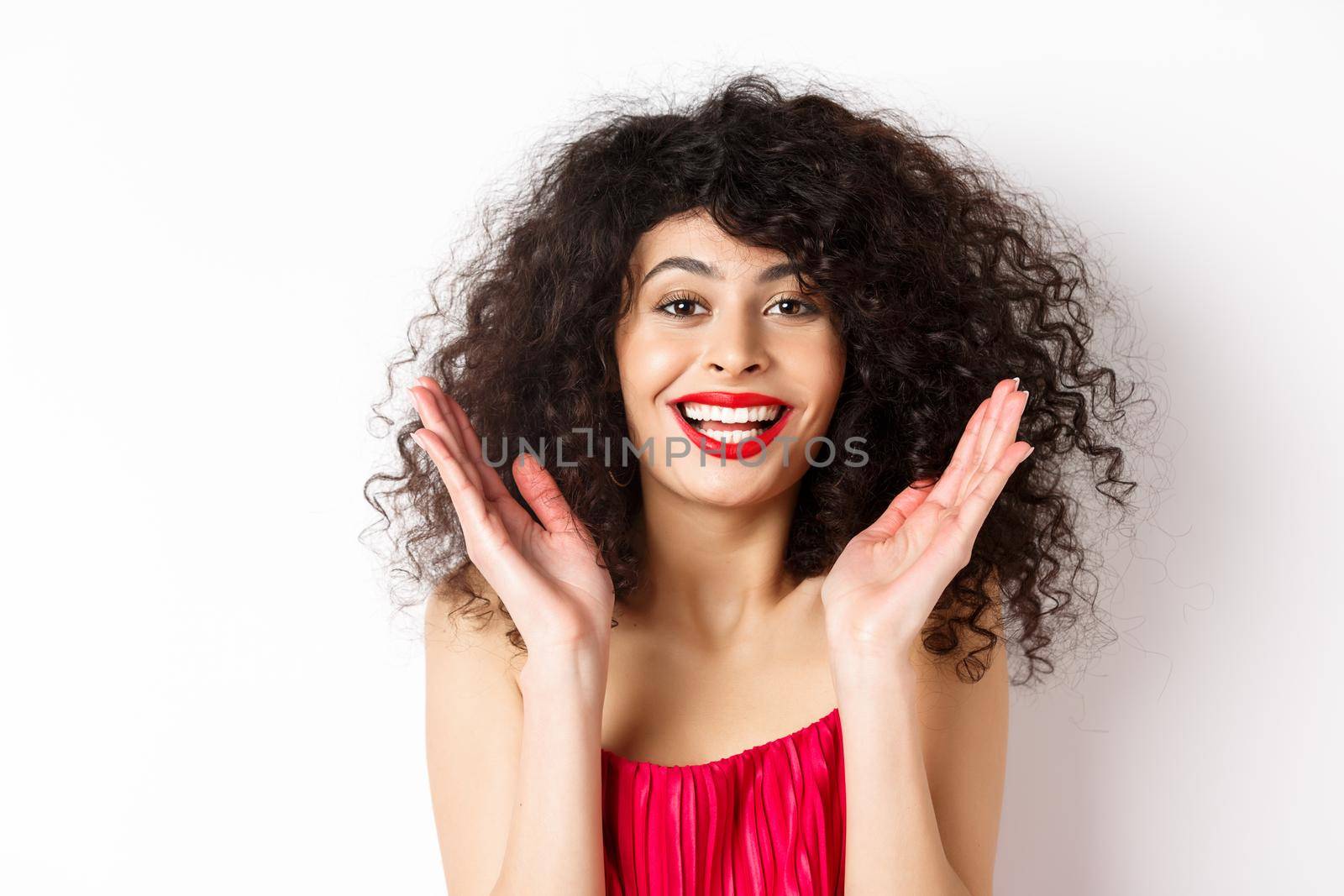 Close-up of fashionable woman with curly hair and red dress, clap hands smiling happy, applause you, standing on white background.