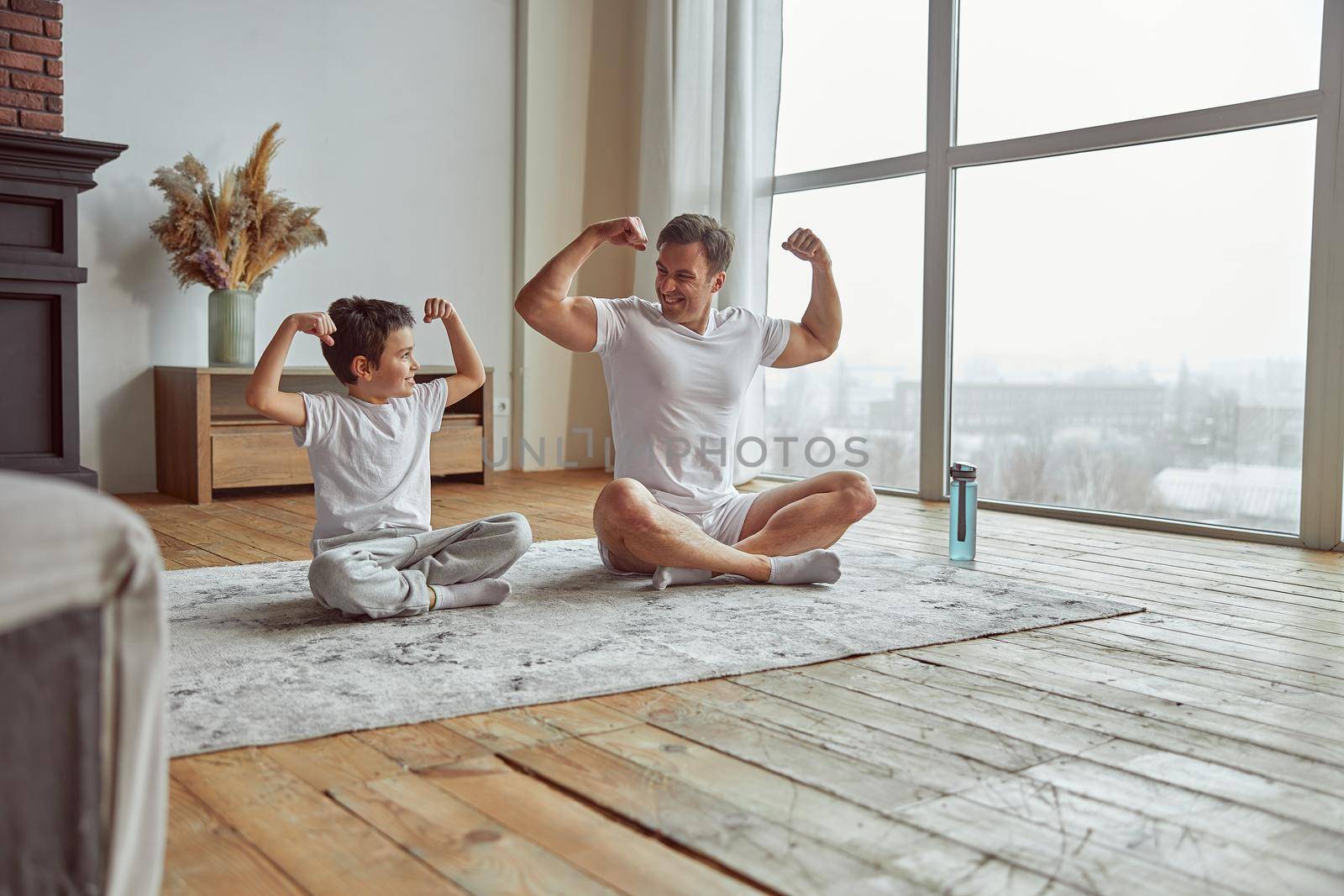 Happy man is sitting with boy on mat and showing biceps after doing workout together