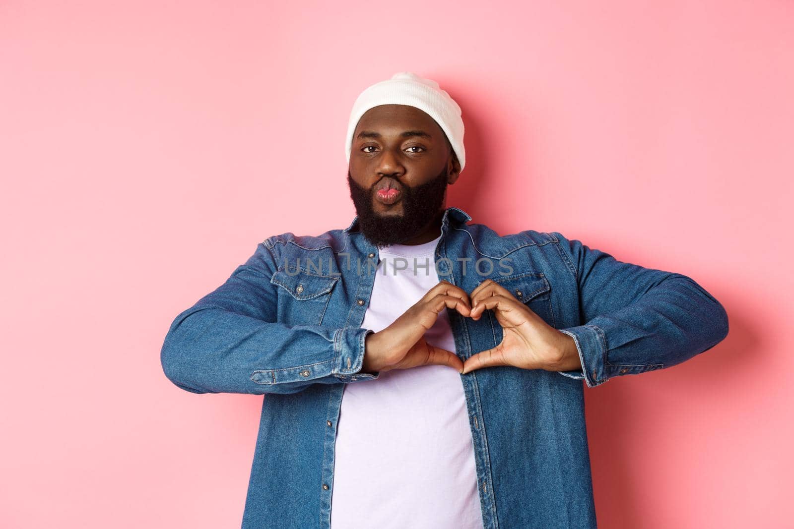 Happy african-american man showing heart sign, I love you gesture, pucker lips for kiss while standing over pink background.