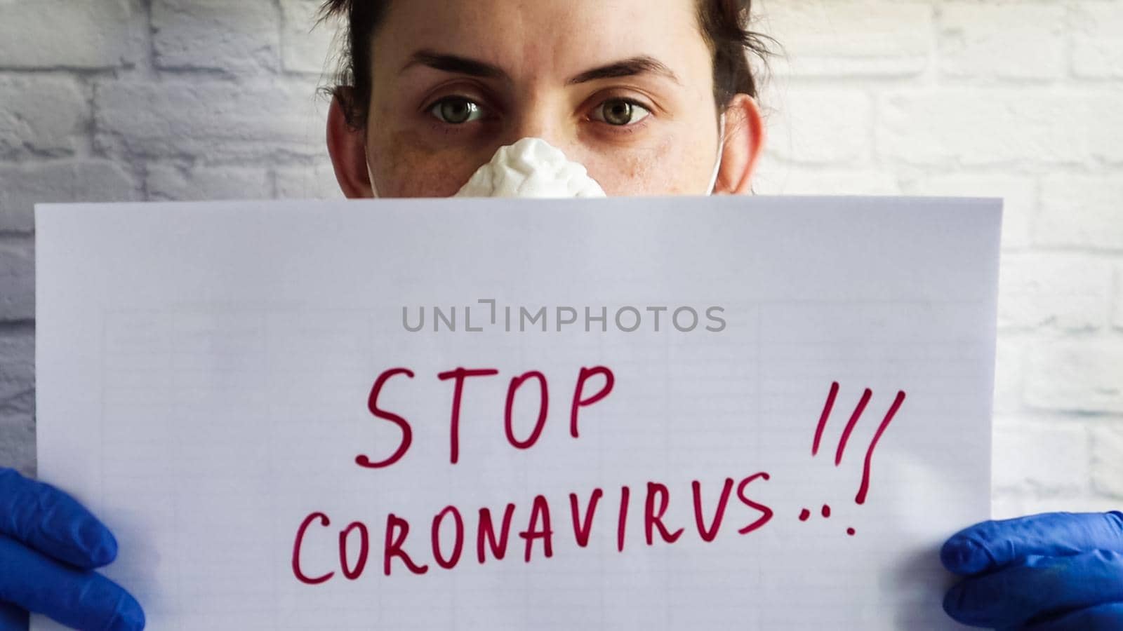 Portrait of a woman in a protective mask that shows the camera close up white sheet of paper with the text stop coronavirus written in red marker. The concept of quarantine coronavirus in the world.