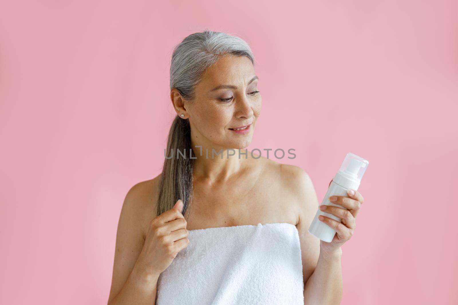 Beautiful mature Asian lady wrapped with towel holds bottle of body care product in studio by Yaroslav_astakhov