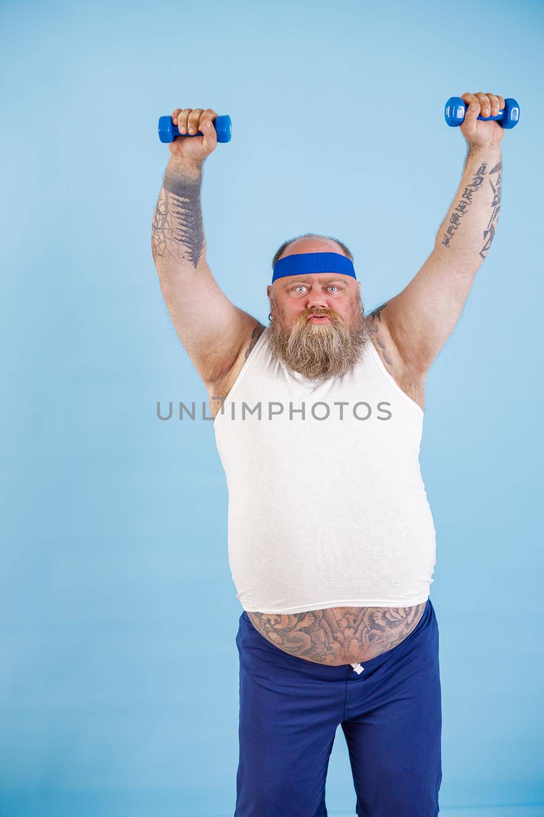 Middle aged fat man with beard does sports exercises with dumbbells trying to loss weight on light blue background in studio