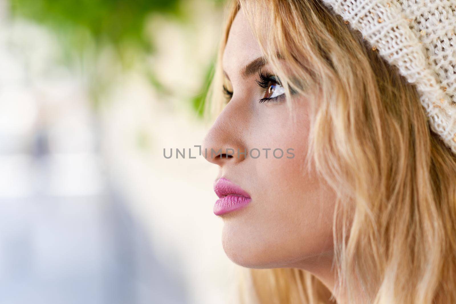 Close-up profile of blonde young woman with curly hair. Girl wearing sweater and wool cap in urban background. Female with beautiful lips painted in pink