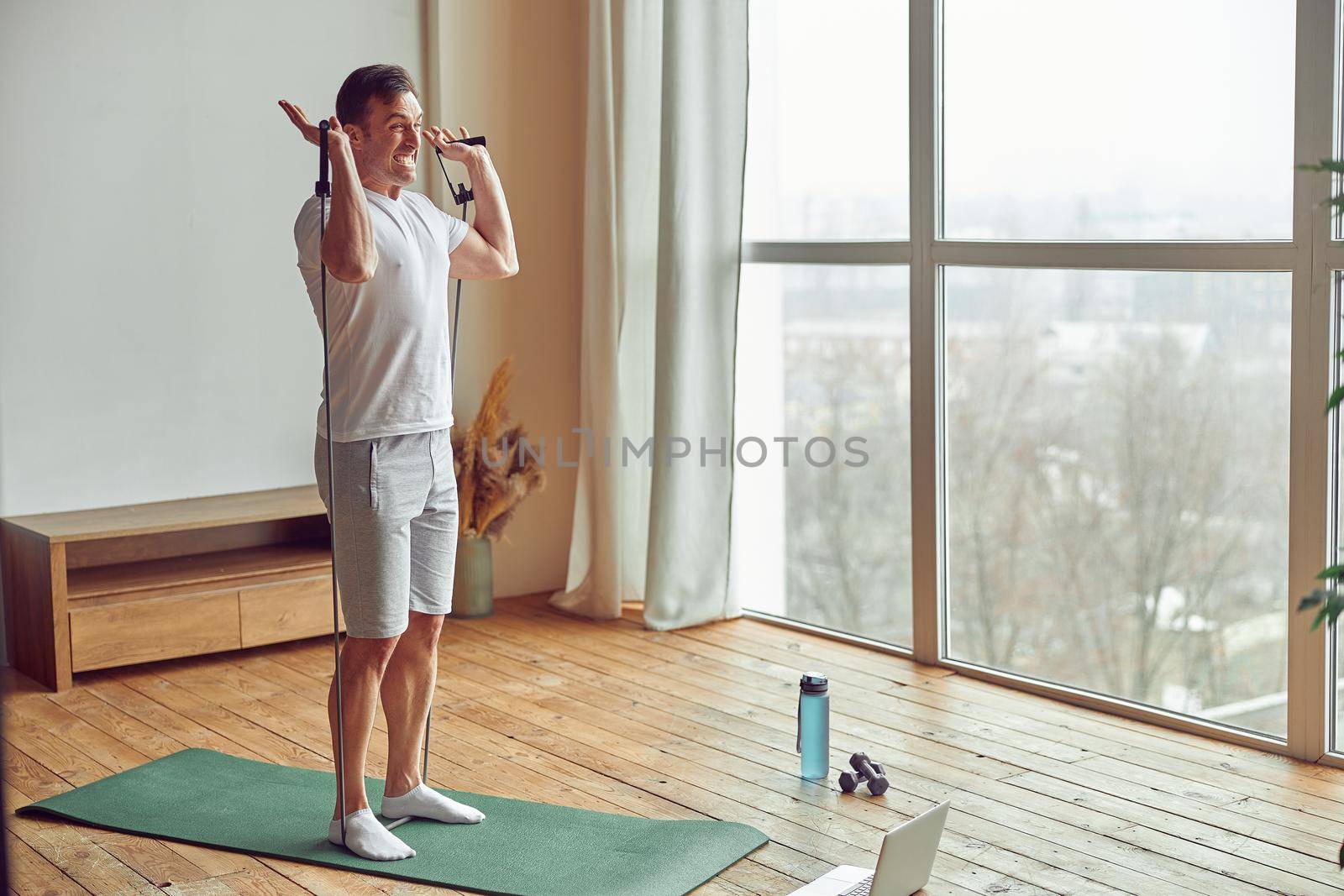 Muscular male is using resistance bands for strength workout at home in front of laptop