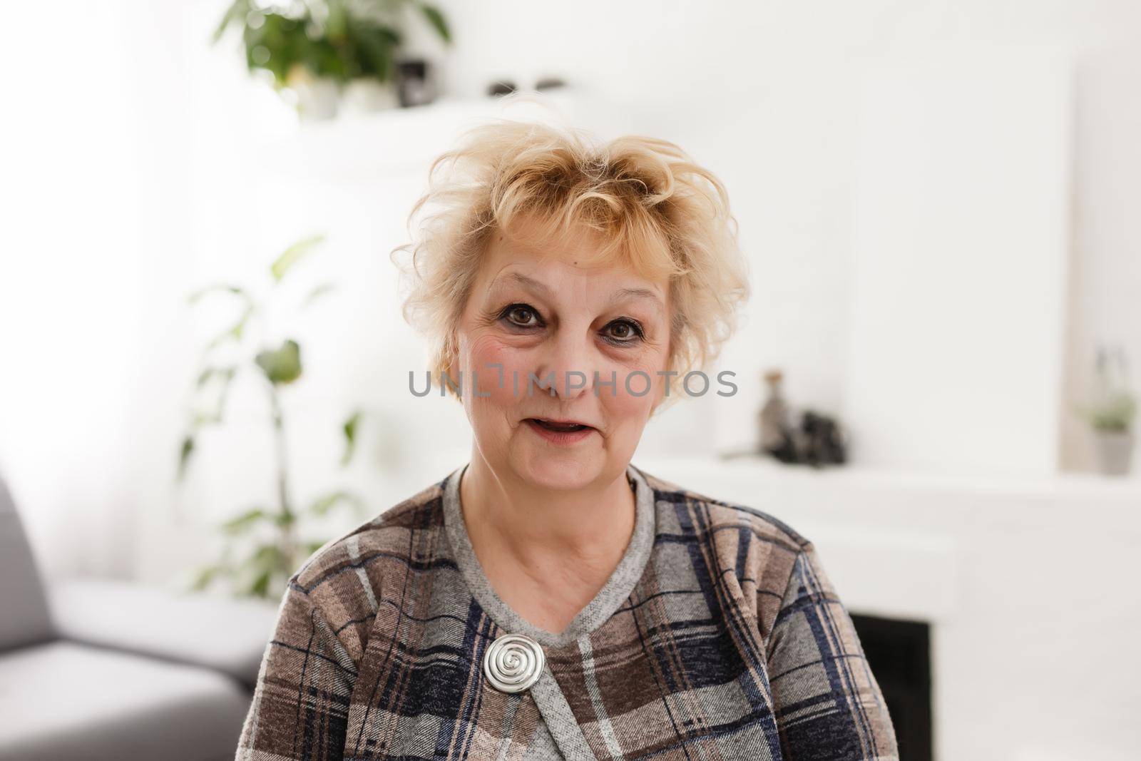 Attractive elderly woman sitting on couch at home, looking at camera, makes video call, communication distantly, modern wireless technologies concept