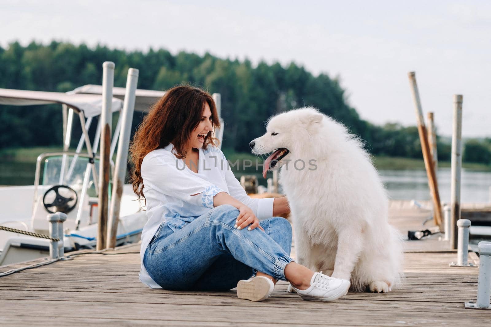 a happy woman with a big white dog sits on a pier by the sea at sunset by Lobachad