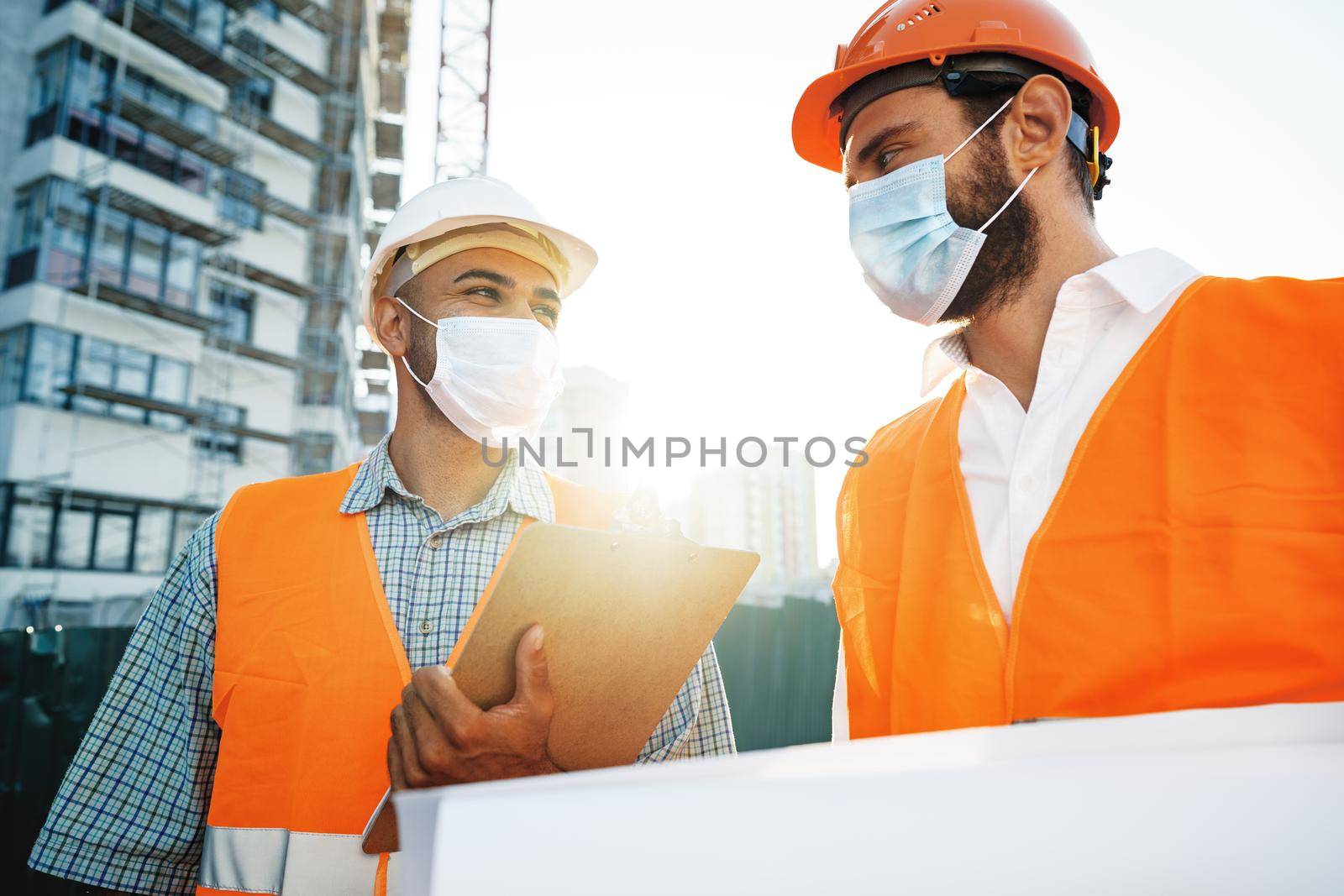 Two men in workwear and medical masks working with blueprints on object, close up