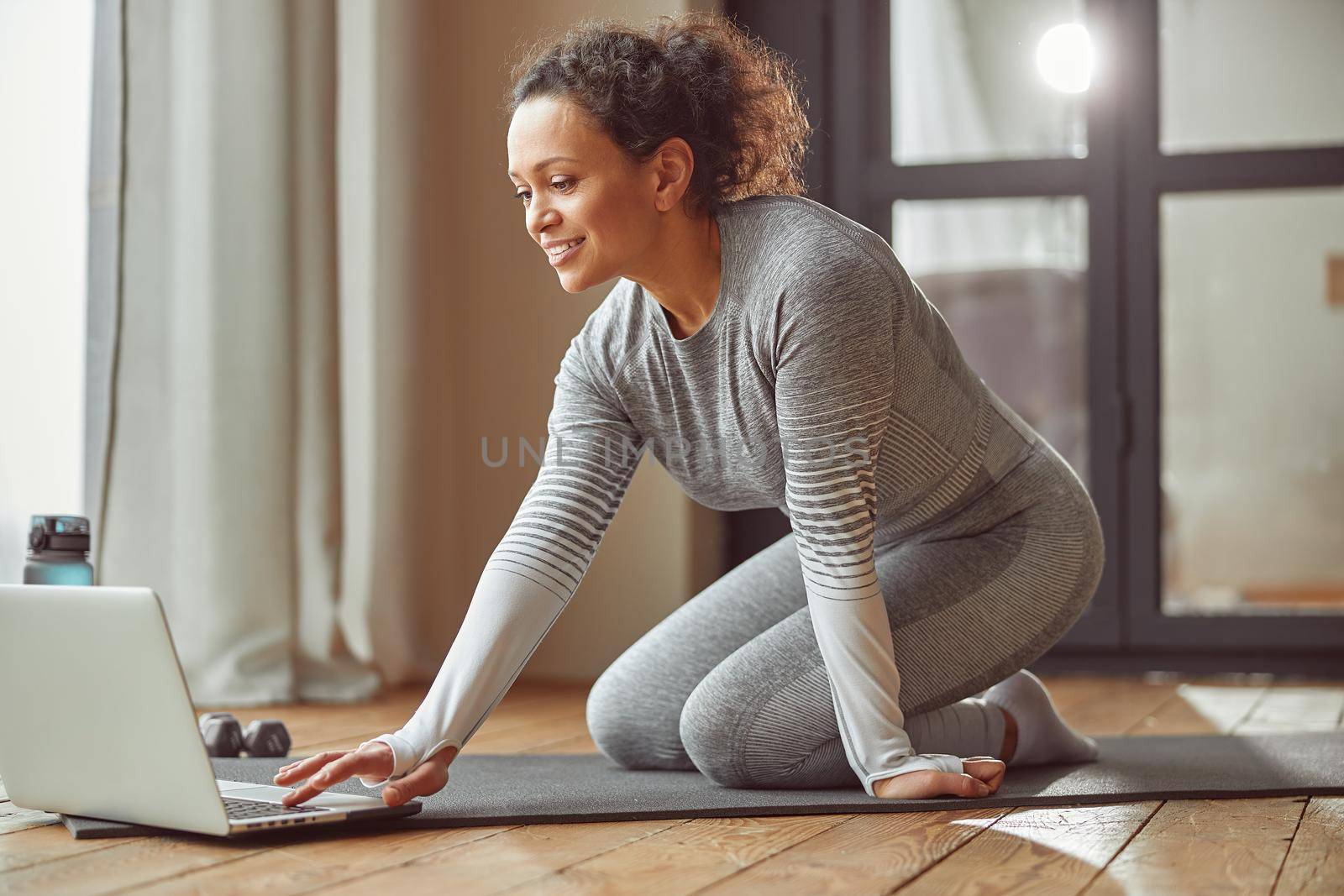 Merry slim woman is sitting on mat and using notebook for online training in living room