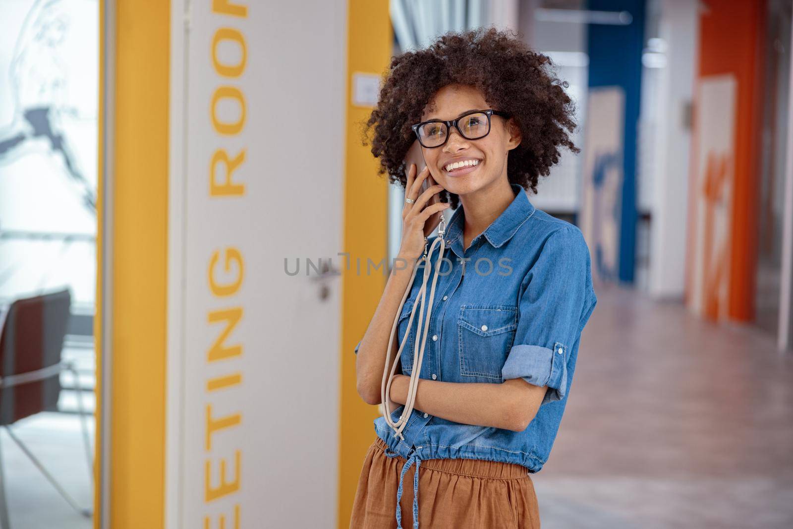 Happy multiethnic lady in glasses standing in office corridor while talking on smartphone while smiling