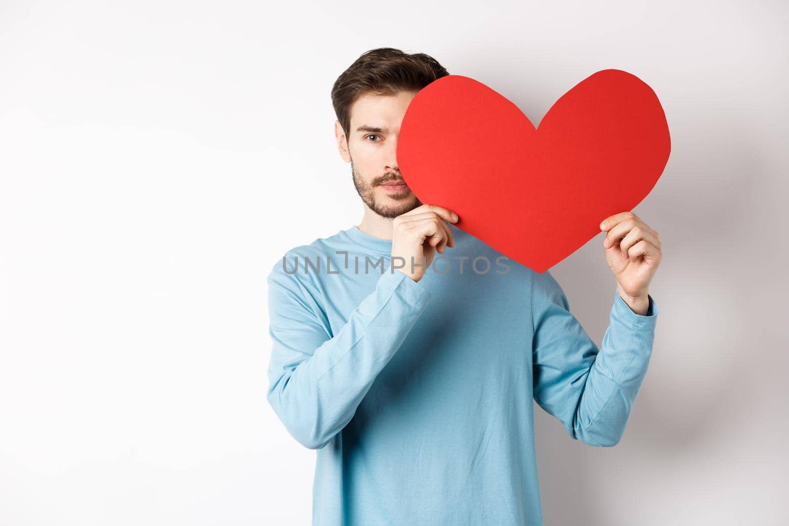 Young caucasian man holding Valentines day romantic heart, cover half of face with cutout and smiling, searching for love date, white background.