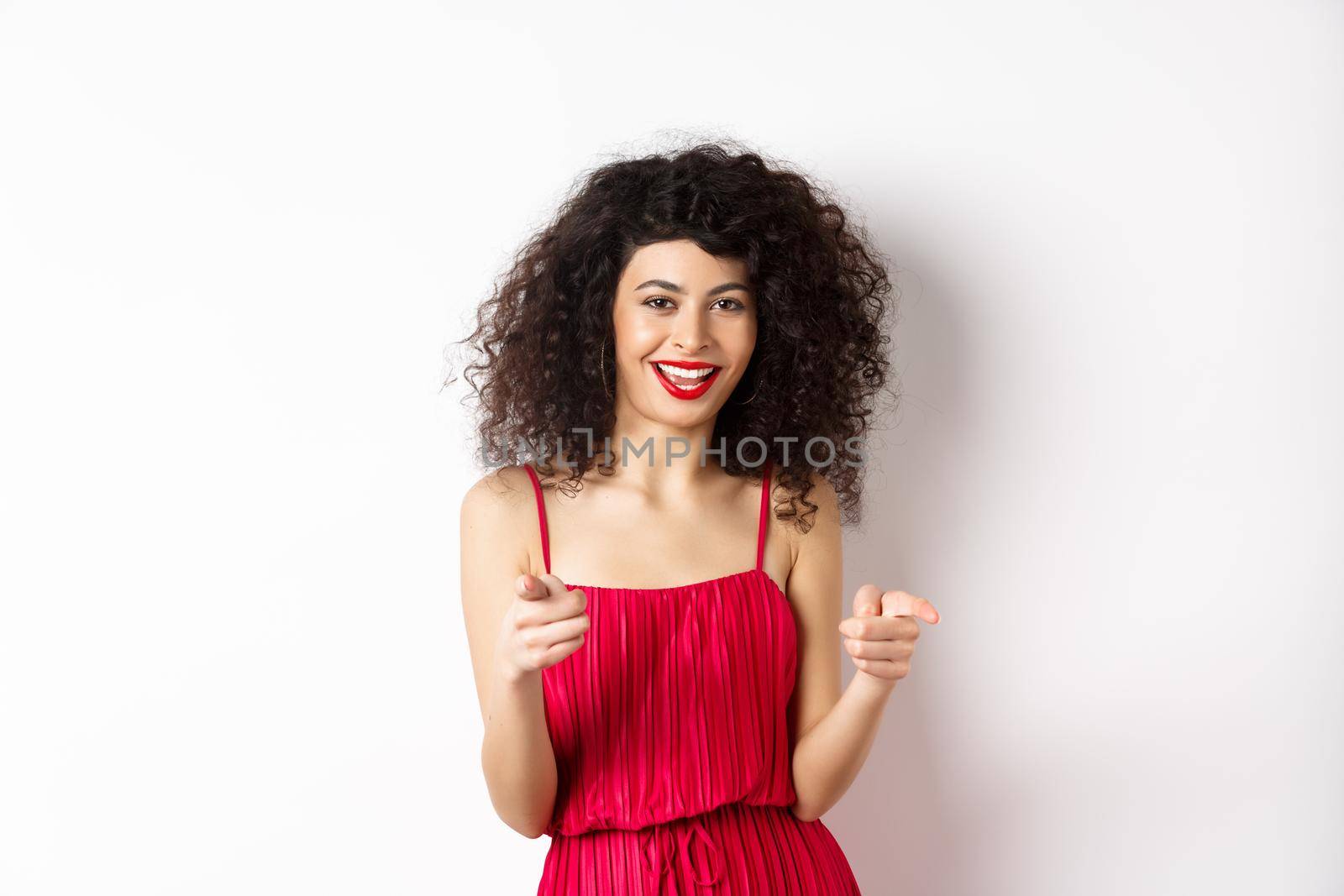 Excited pretty woman in red dress congratulate you, pointing fingers at camera and smiling, standing on white background.
