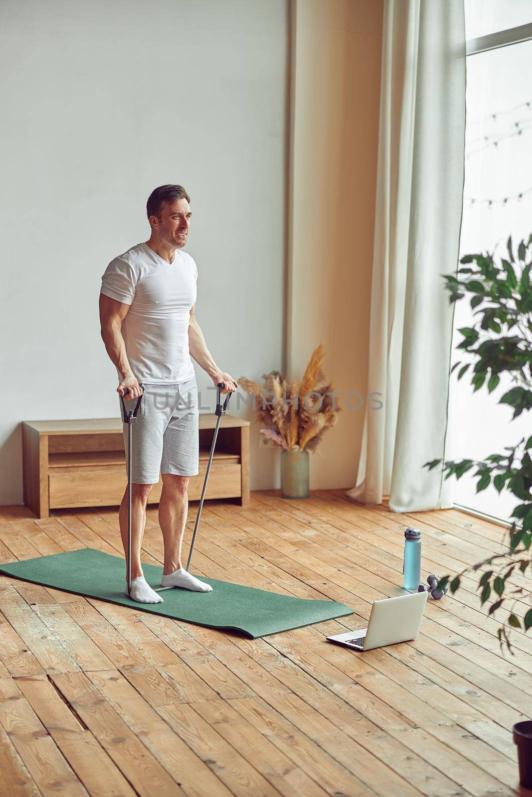 Athletic man is using resistance bands for doing arms workout in front of laptop near large window