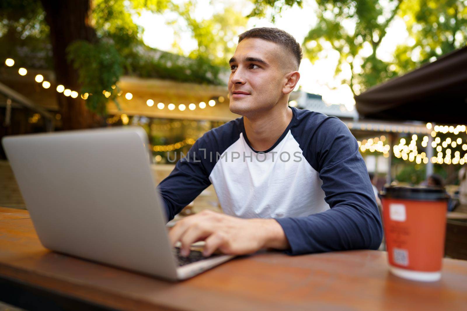 Young man sitting at table and typing on laptop keyboard while working in outdoor cafe by Fabrikasimf