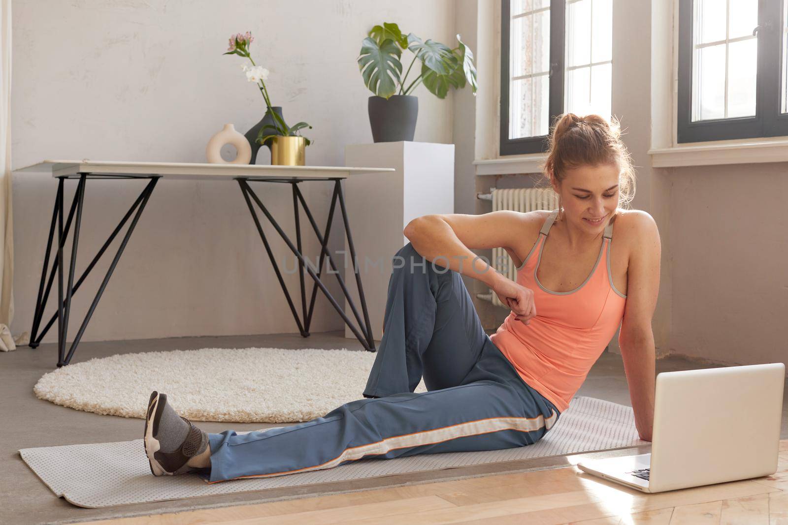 Smiling athletic female in sportswear sitting on mat with laptop during online workout at home and looking at screen