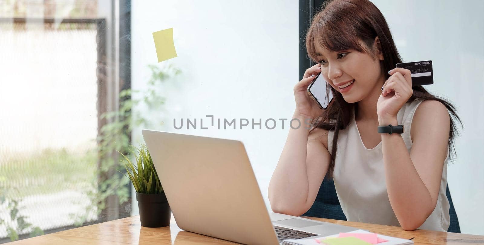 Smiling woman sitting in office talking on mobile phone and paying online by credit card on tablet computer.