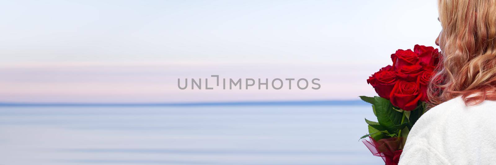 Happy woman in white robe with a bouquet of red roses looks from the hotel balcony at the sea by PhotoTime