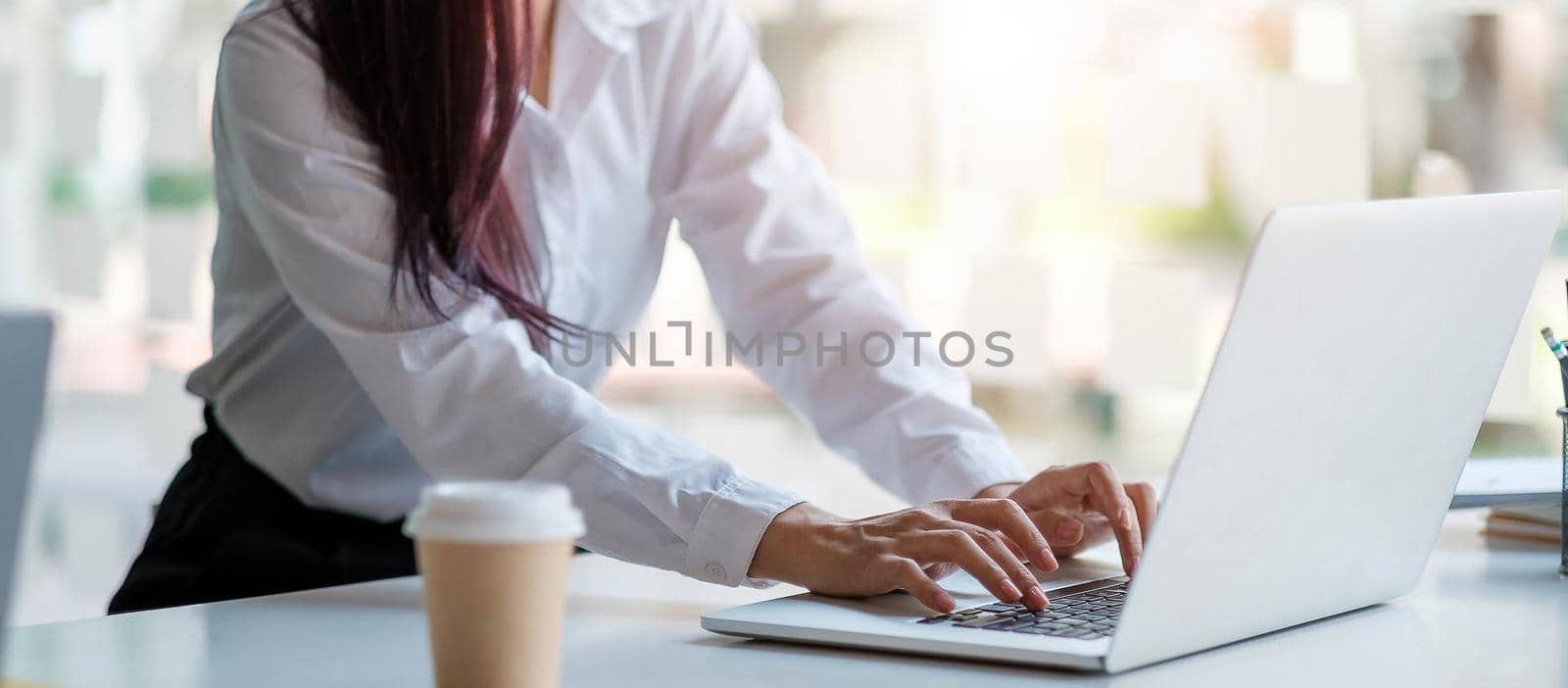 Close up of tanned woman's hands on a computer keyboard. She is typing. There is a piece of paper with blue graphs and a cup of coffee.