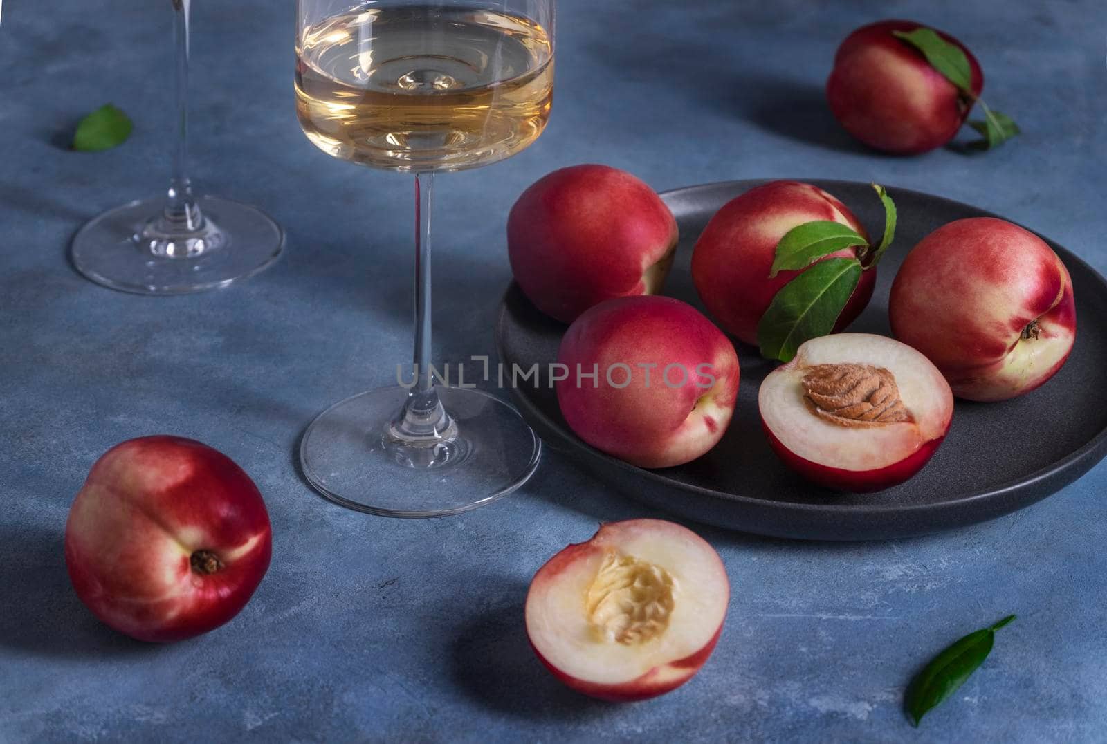 Several ripe peaches or nectarines on a black ceramic plate and a glass of white wine sit on a blue plaster-textured surface. Selective focus.