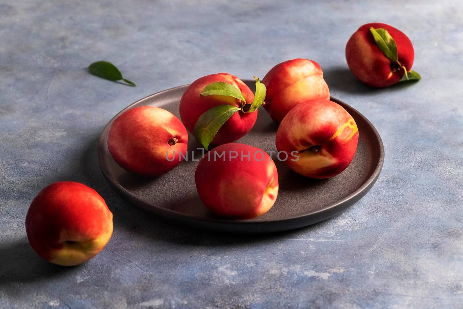 Several ripe peaches or nectarines lie on a black ceramic plate on a blue plaster-textured surface. Selective focus.