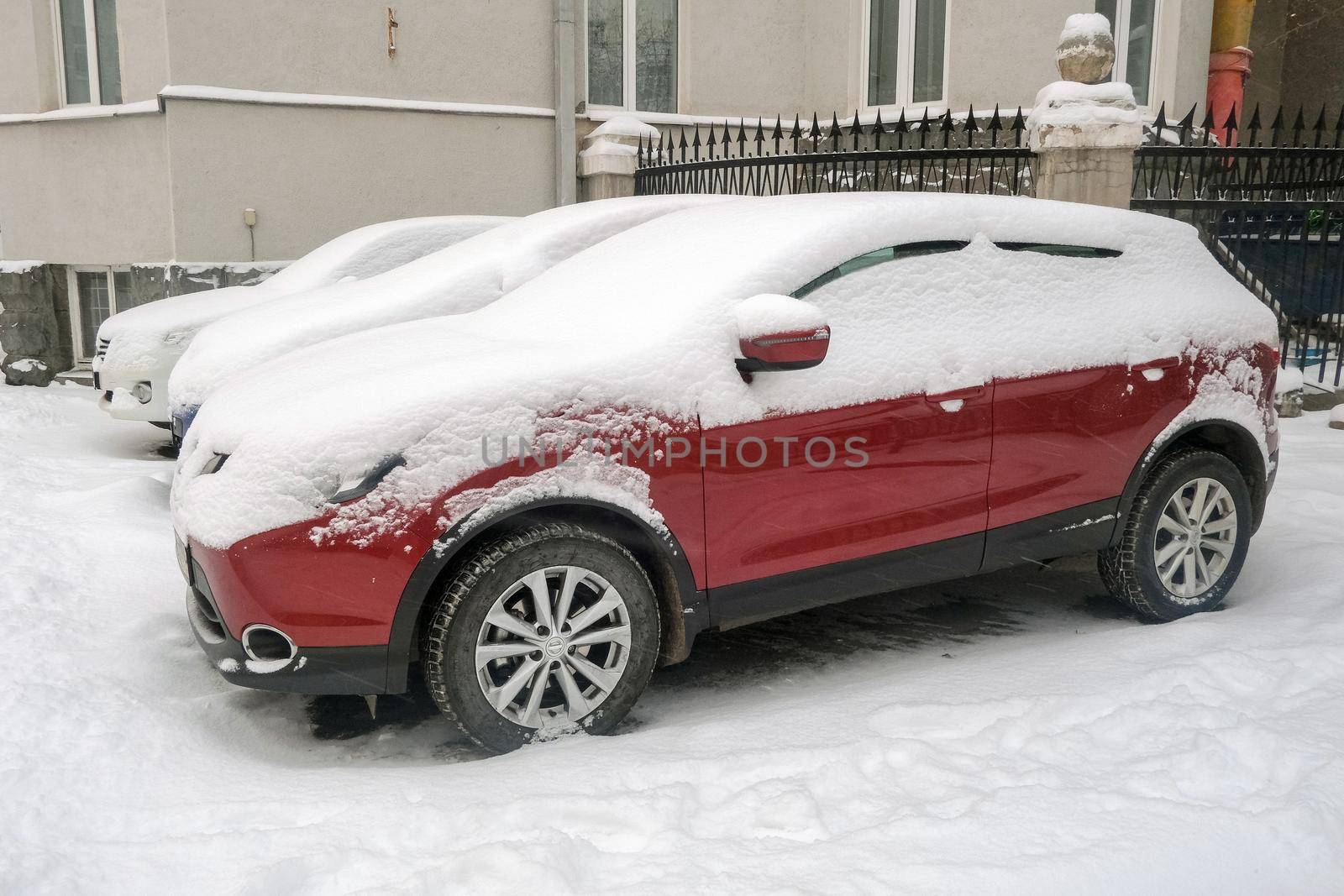 Modern red automobile covered with white snow parked near house on street in wintertime