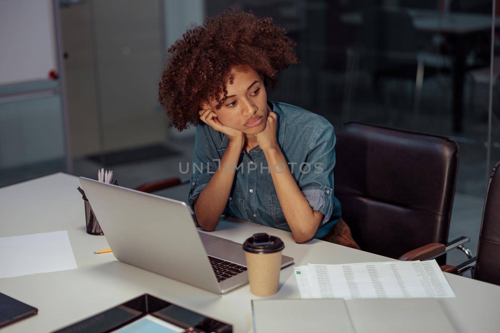 Upset multiethnic lady working in the office while sitting at desk with laptop and cup of coffee