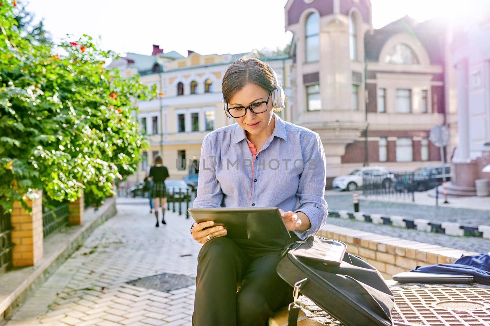 Serious mature business woman in glasses headphones with digital tablet listening attentively and looking at tablet screen, confident female sitting on city street in sunset light