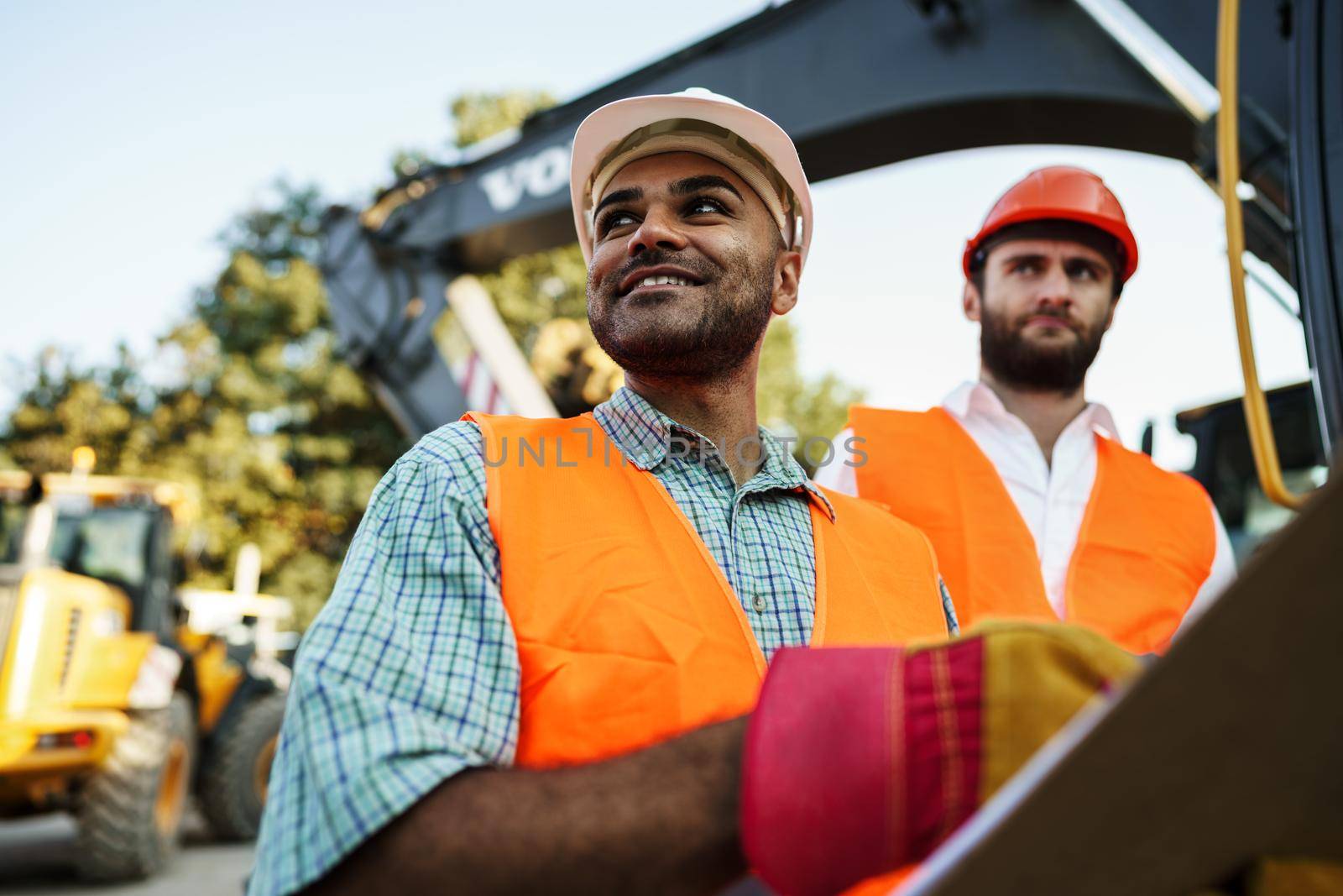 Two men engineers in workwear discussing their work standing against construction machines