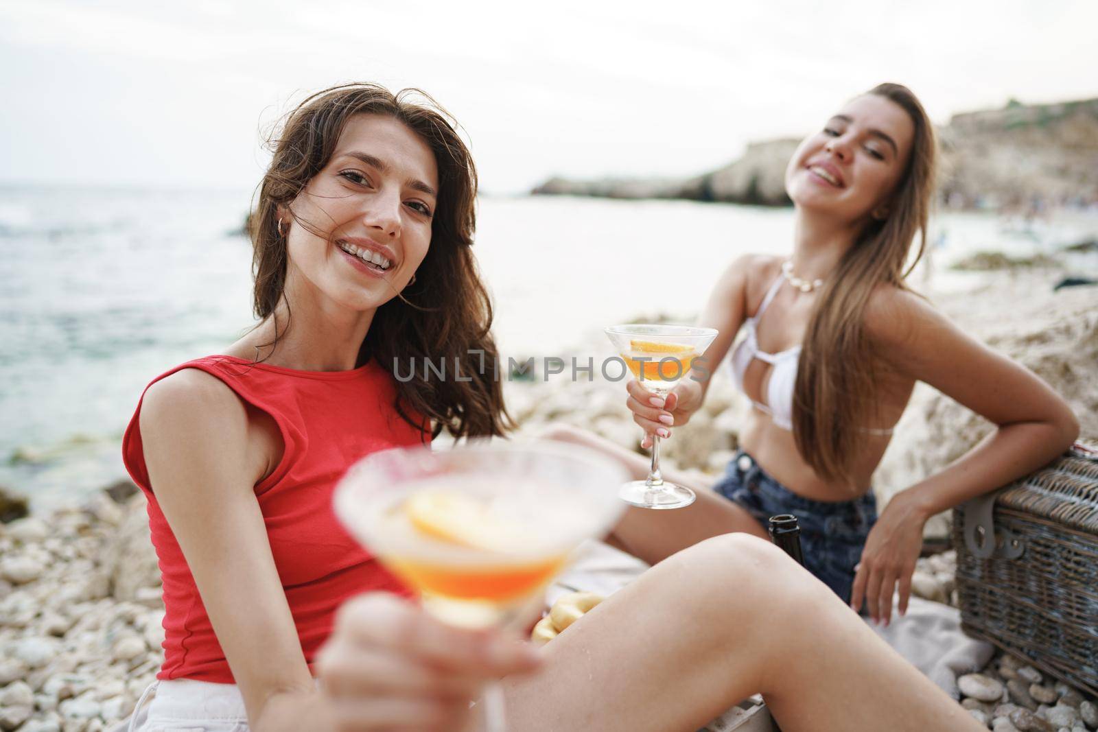 Two young female friends having a picnic on a beach drinking cocktails