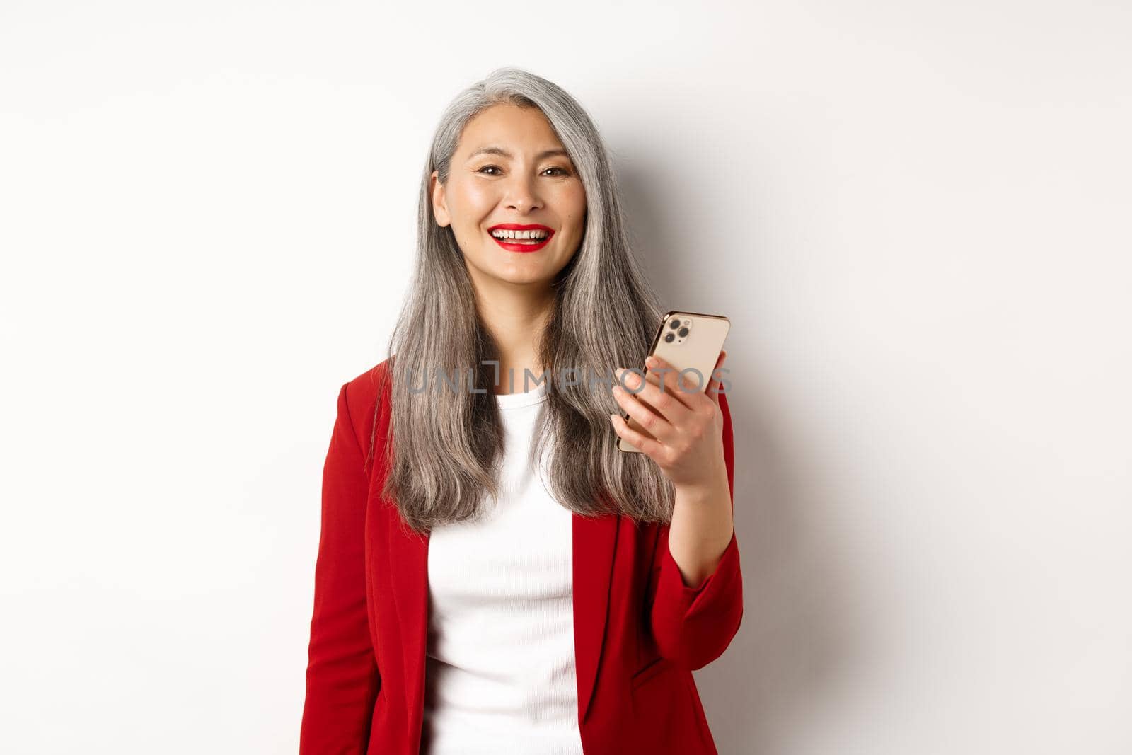 Asian female entrepreneur in red blazer using smartphone, smiling happy at camera, standing over white background by Benzoix
