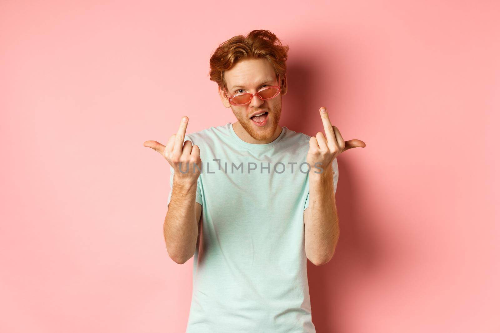 Arrogant and careless redhead man in sunglasses dont give a fuck, showing middle fingers at camera and frowning, standing over pink background by Benzoix