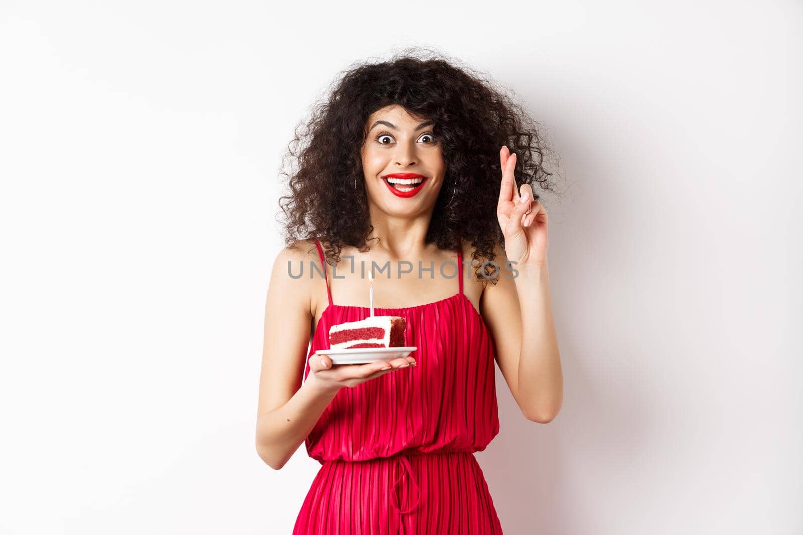 Excited birthday girl in red dress, cross fingers while making wish and blowing candle on bday cake, smiling happy, white background.