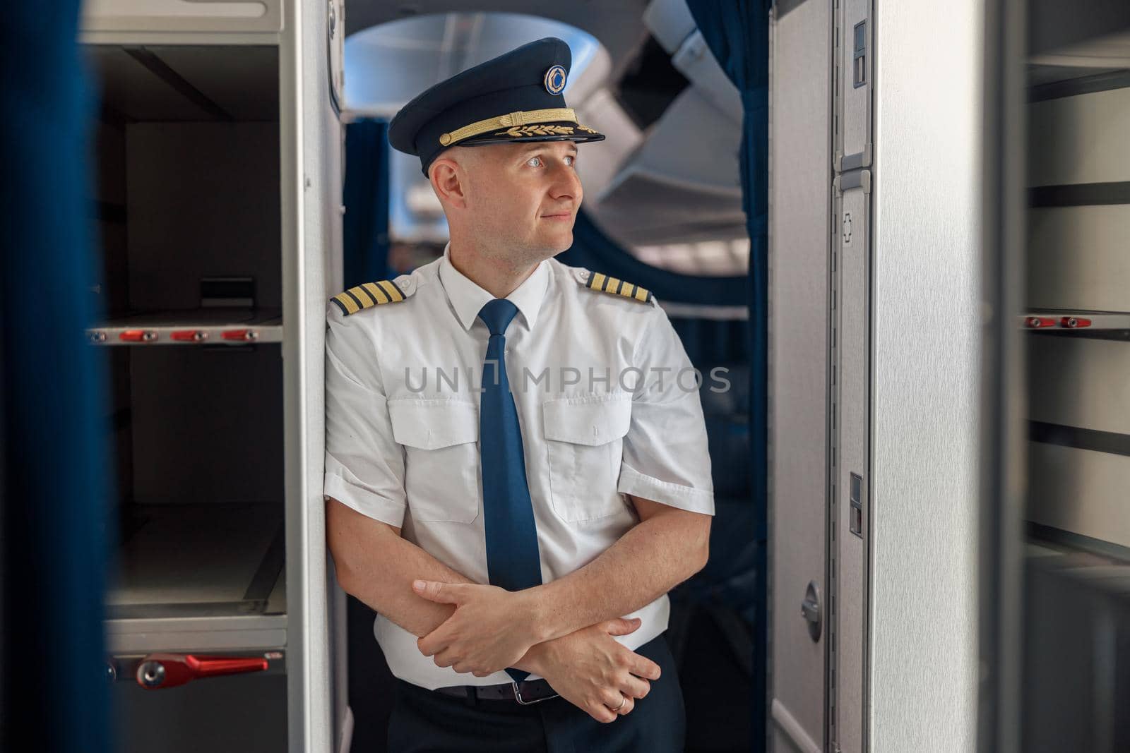 Thoughtful male pilot in uniform and hat keeping arms crossed and looking aside while standing inside of the airplane. Transportation, aircrew concept