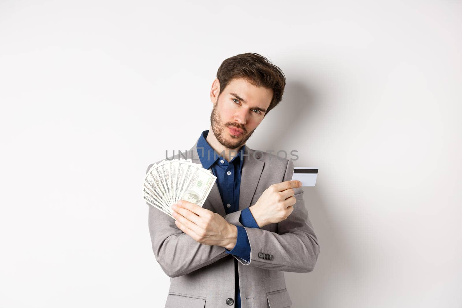 Cool young rich man in suit showing dollar bills and plastic credit card, making money, standing on white background.