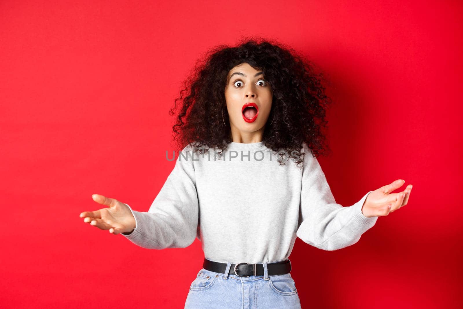 Beautiful young woman standing in awe, drop jaw and looking impressed at camera, spread hands sideways with disbelief, standing on red background.