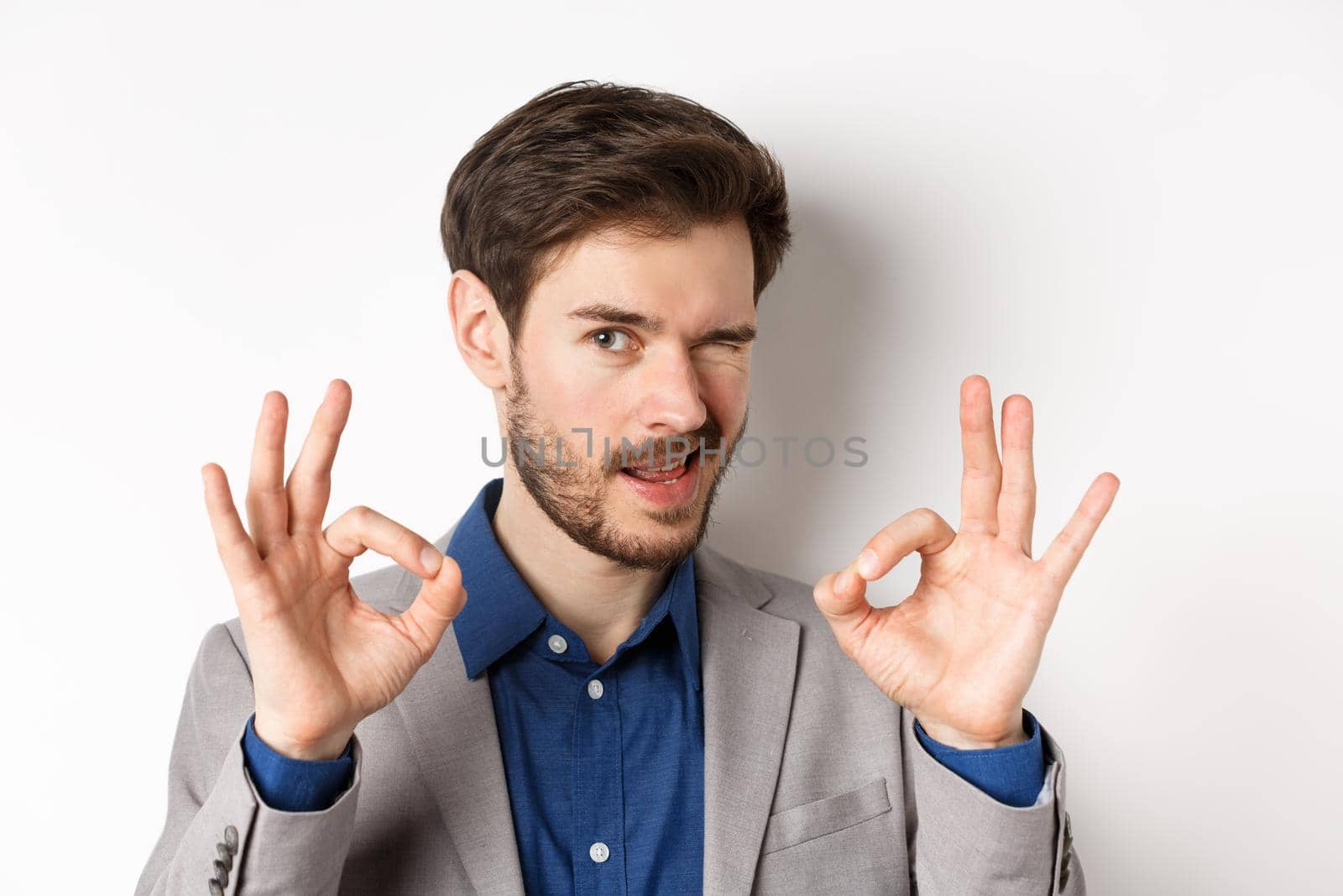 Close up of confident handsome man assuring all good, showing okay signs and winking at camera, no problem gesture, standing on white background.