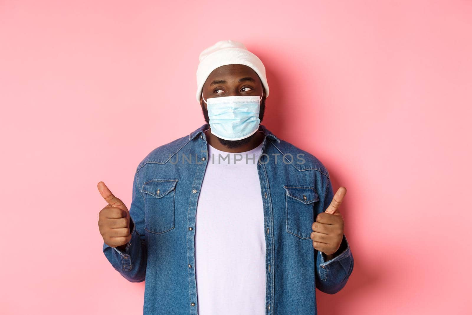 Coronavirus, lifestyle and social distancing concept. Black hipster guy in beanie and medical mask showing thumbs-up, praising product, looking left and smiling, pink background.