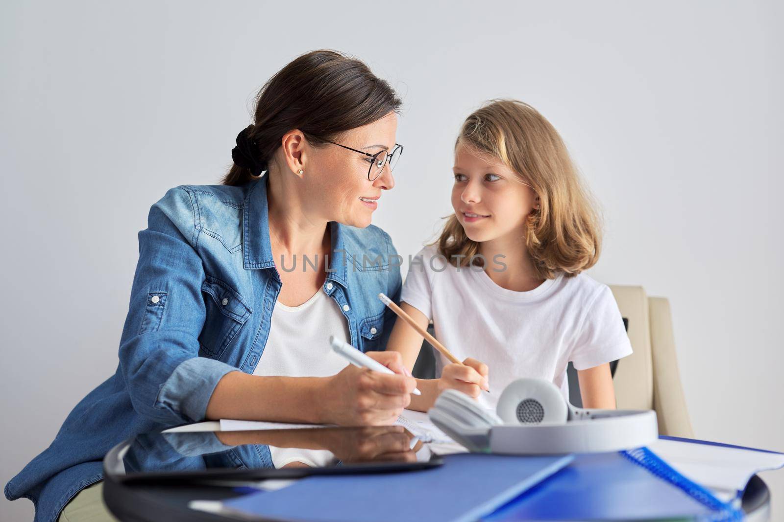 Mom and daughter student learning school lessons together at home, parent helping child, using digital tablet, lessons on Internet, girl in headphones, writes in notebook with pencil