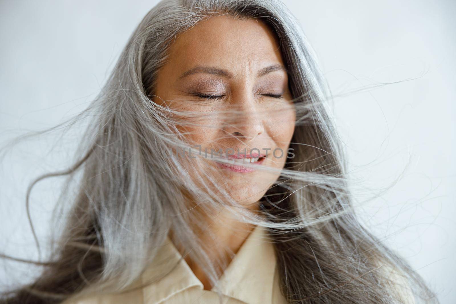 Happy mature Asian model with flying hoary hair stands on light grey background by Yaroslav_astakhov