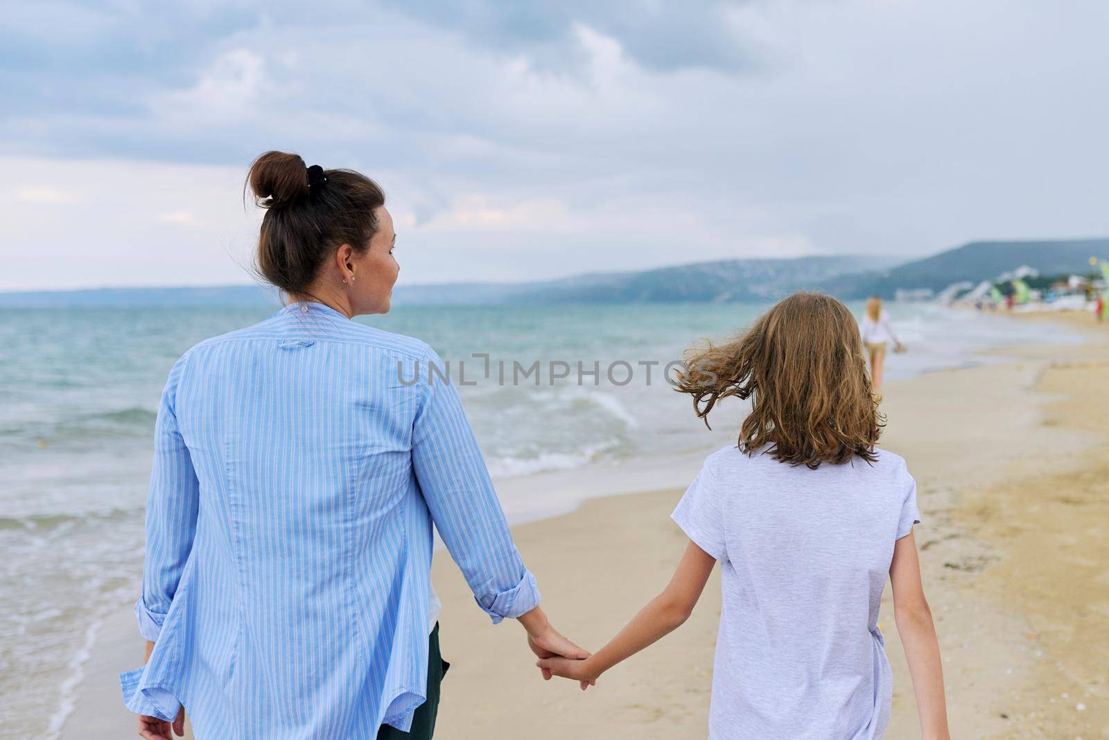 Happy mom and daughter walking on the sea beach holding hands, back view. Family middle-aged mother and preteen child together, vacation, sea weekend, travel, love, happiness and joy
