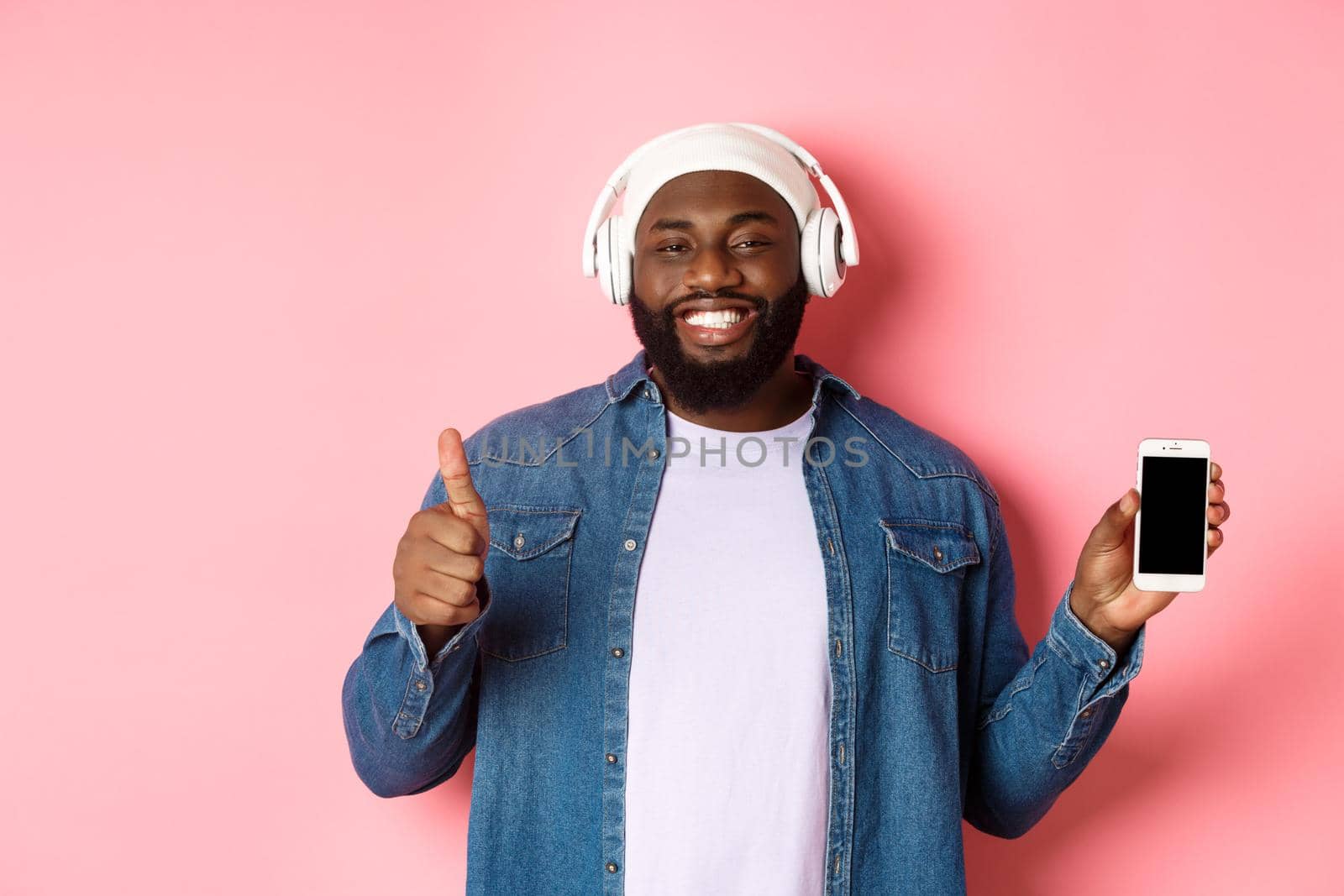 Happy hipster guy listening music on headphones and showing mobile screen, smiling satisfied, showing thumb-up in approval, like playlist, standing over pink background.