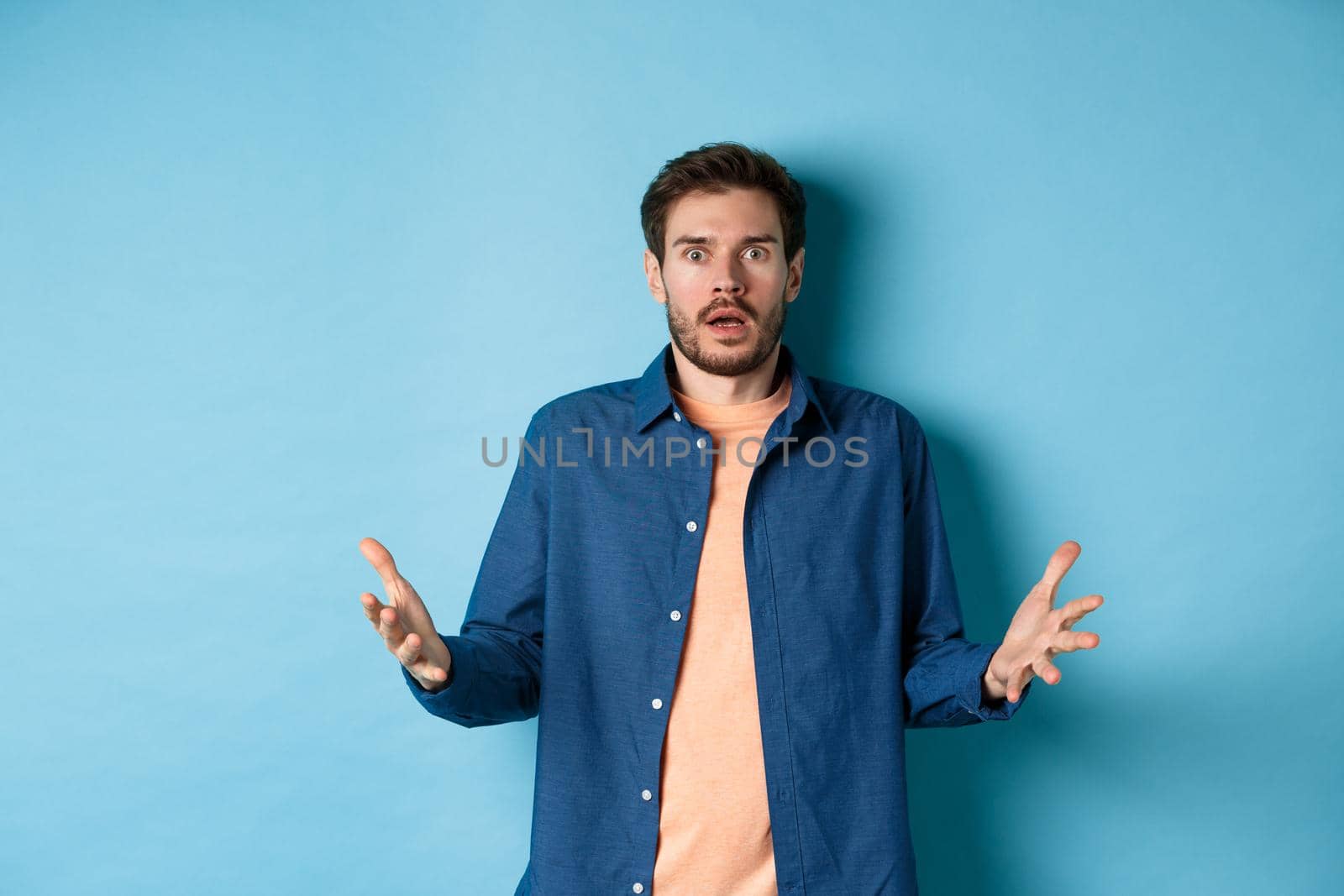 Shocked young man raising hands up and staring at camera startled, look with disbelief, standing on blue background by Benzoix