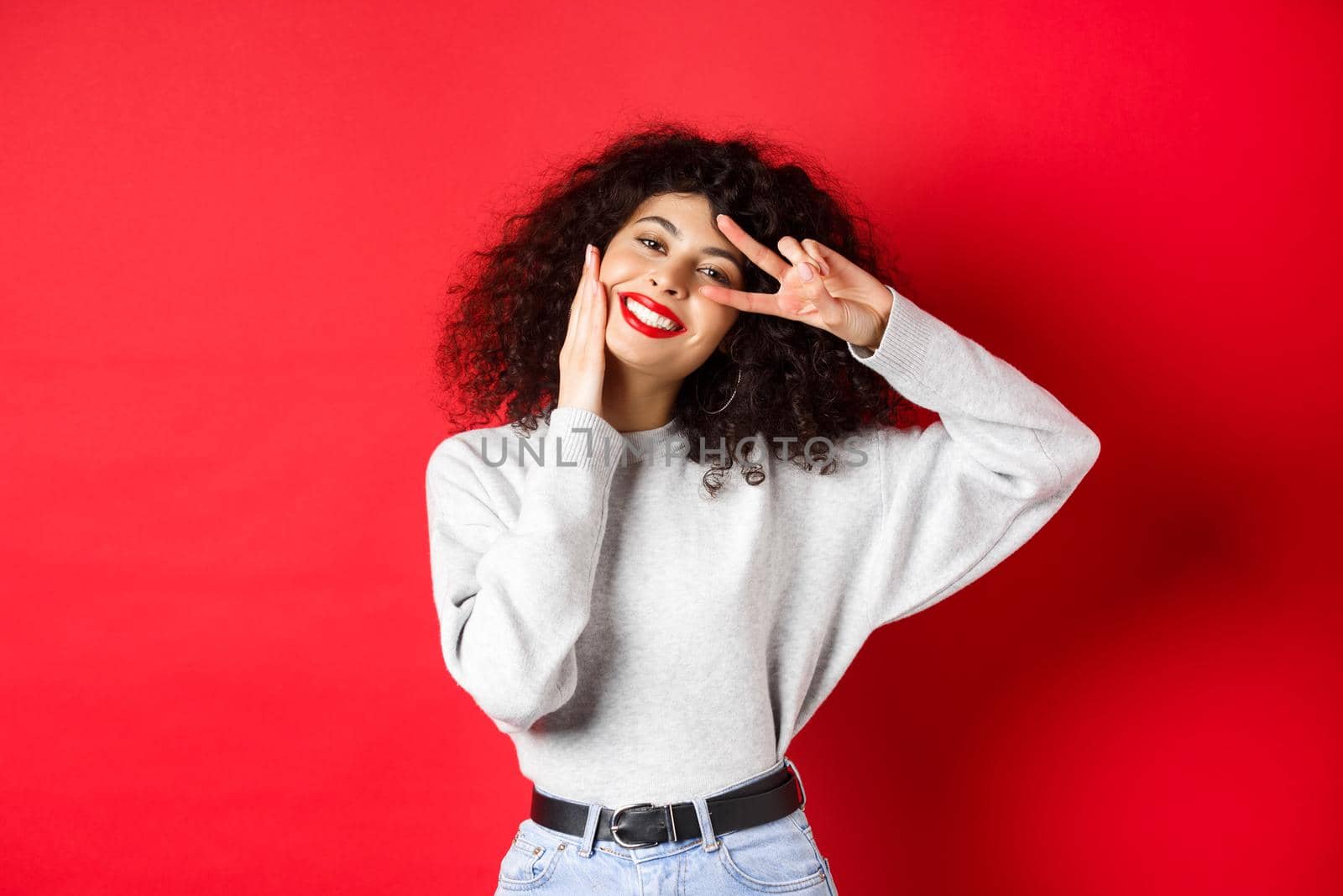 Beauty and makeup. Happy young woman with curly hair, touching face and showing v-sign with cute smile, standing against red background by Benzoix