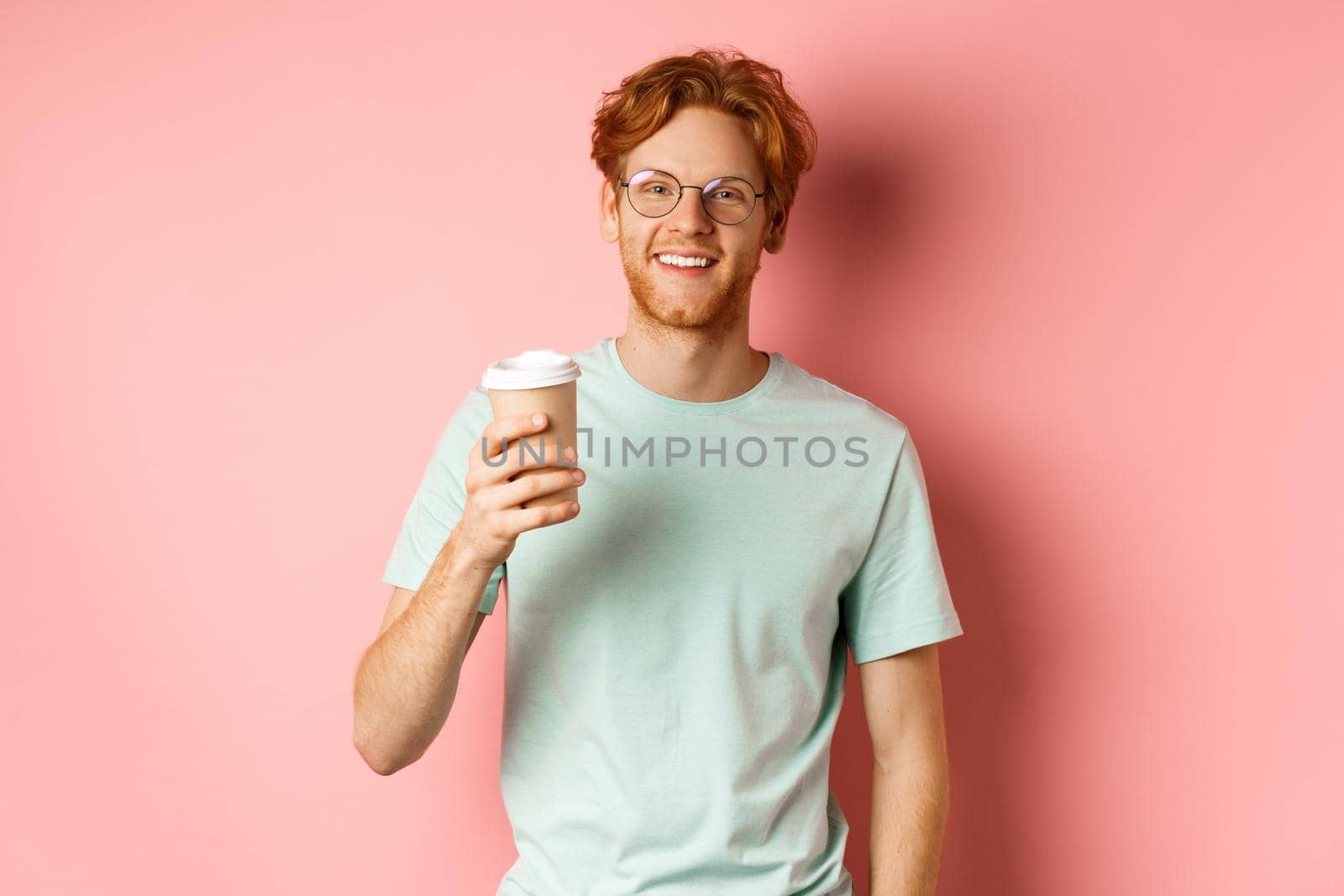 Happy redhead man in glasses and t-shirt drinking coffee and smiling, enjoying lunch break, holding takeaway cup, standing over pink background by Benzoix