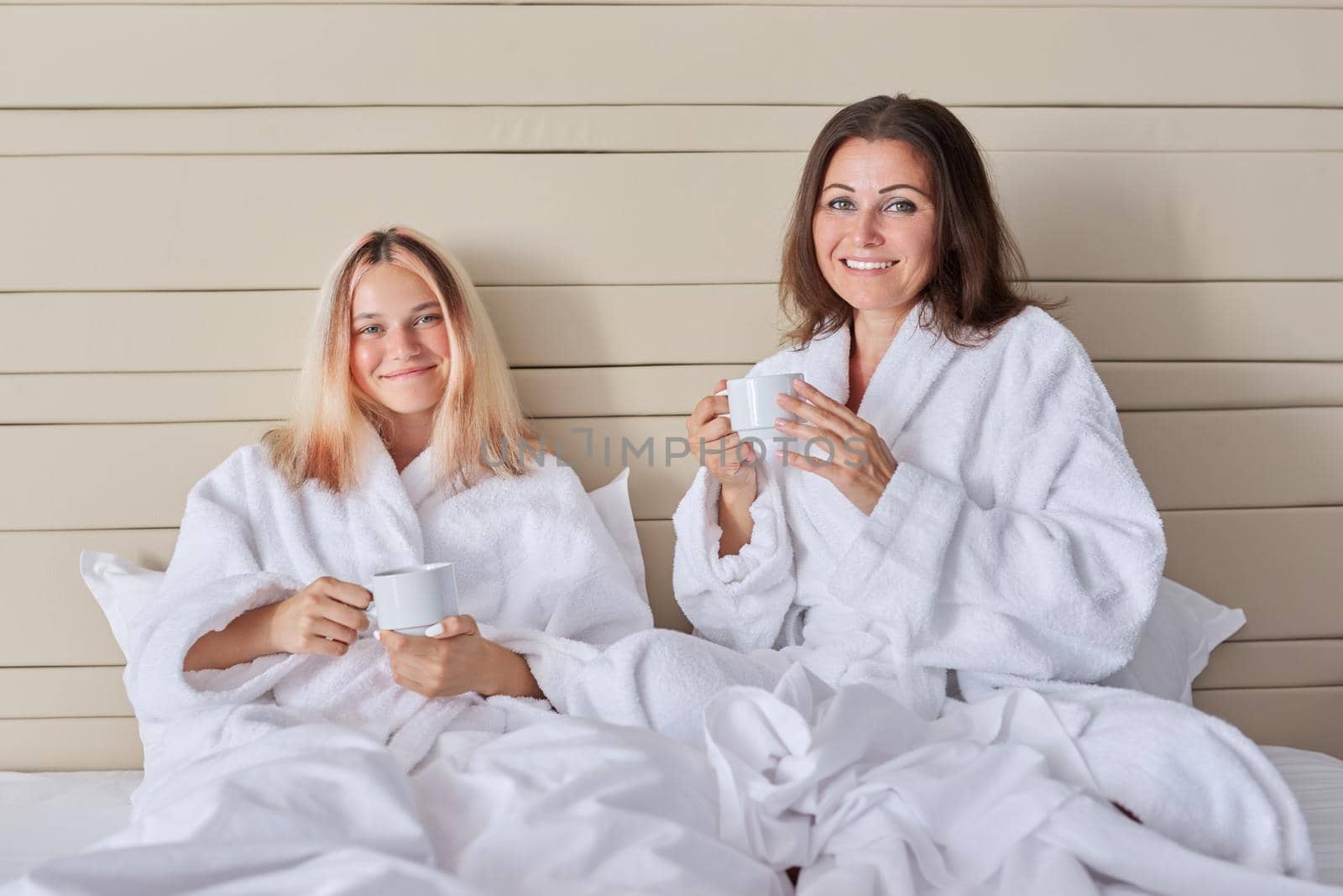 Happy mother and teenage daughter drinking coffee talking. Women in white bathrobes, resting sitting in bed, vacation together. Communication between parent and adolescent child, family, relationship