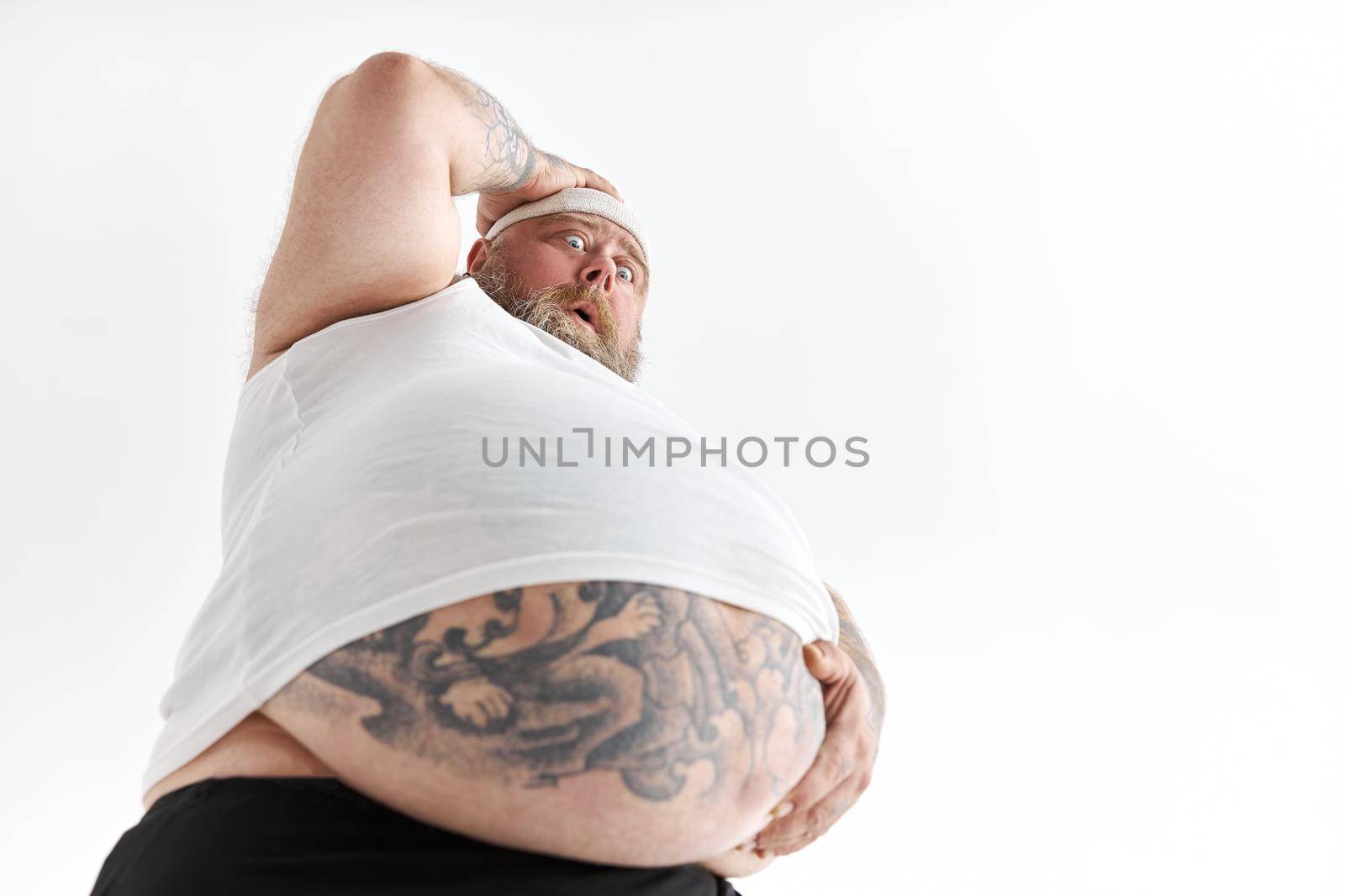 fat man with big belly and tattoes in sports wear is holding his stomach with shocked emotion