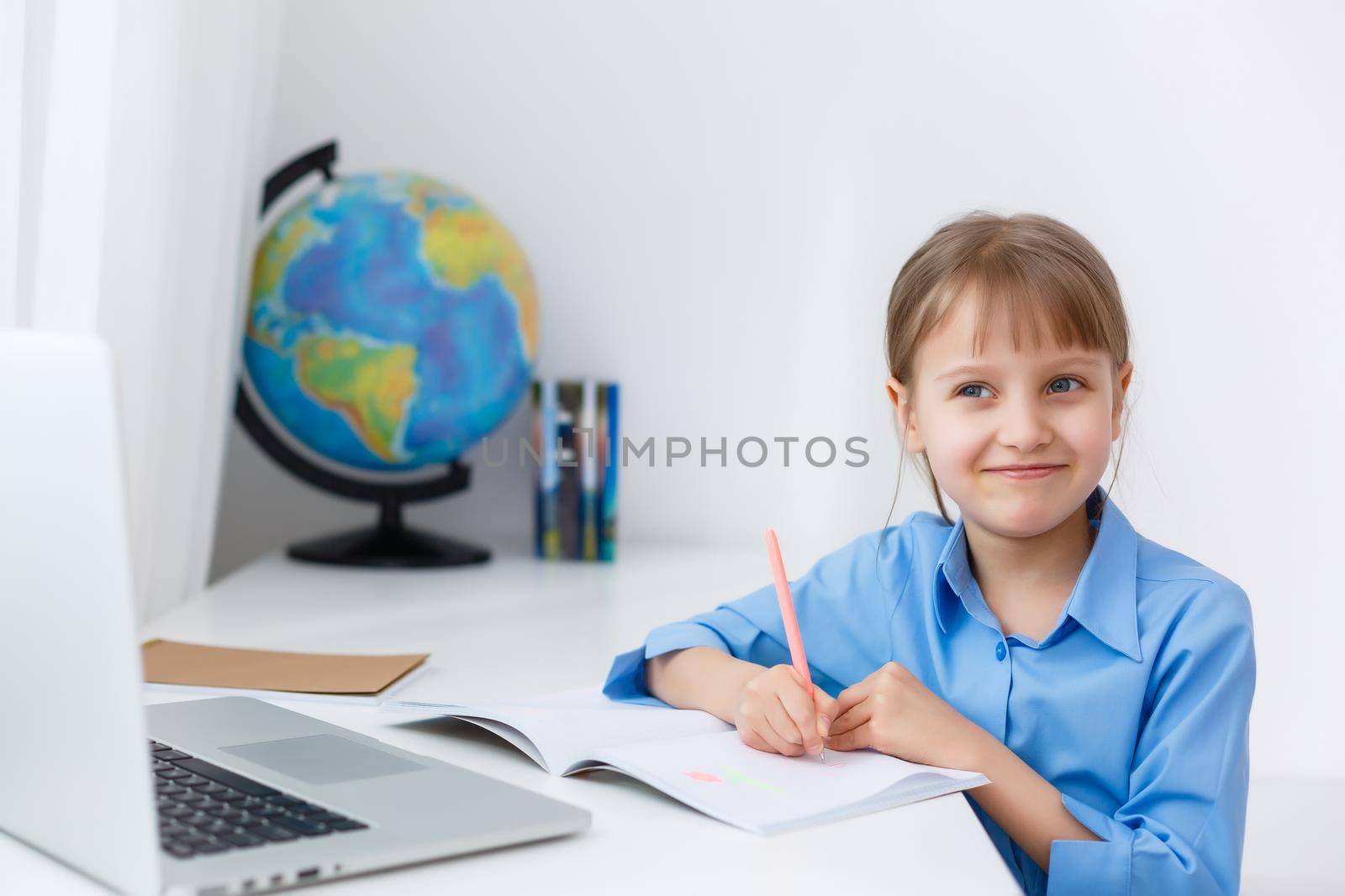 Smart Little Girl Does Homework in Her Living Room. She's Sitting at Her Desk Writes with a Pen in Her Textbooks and Uses Laptop.